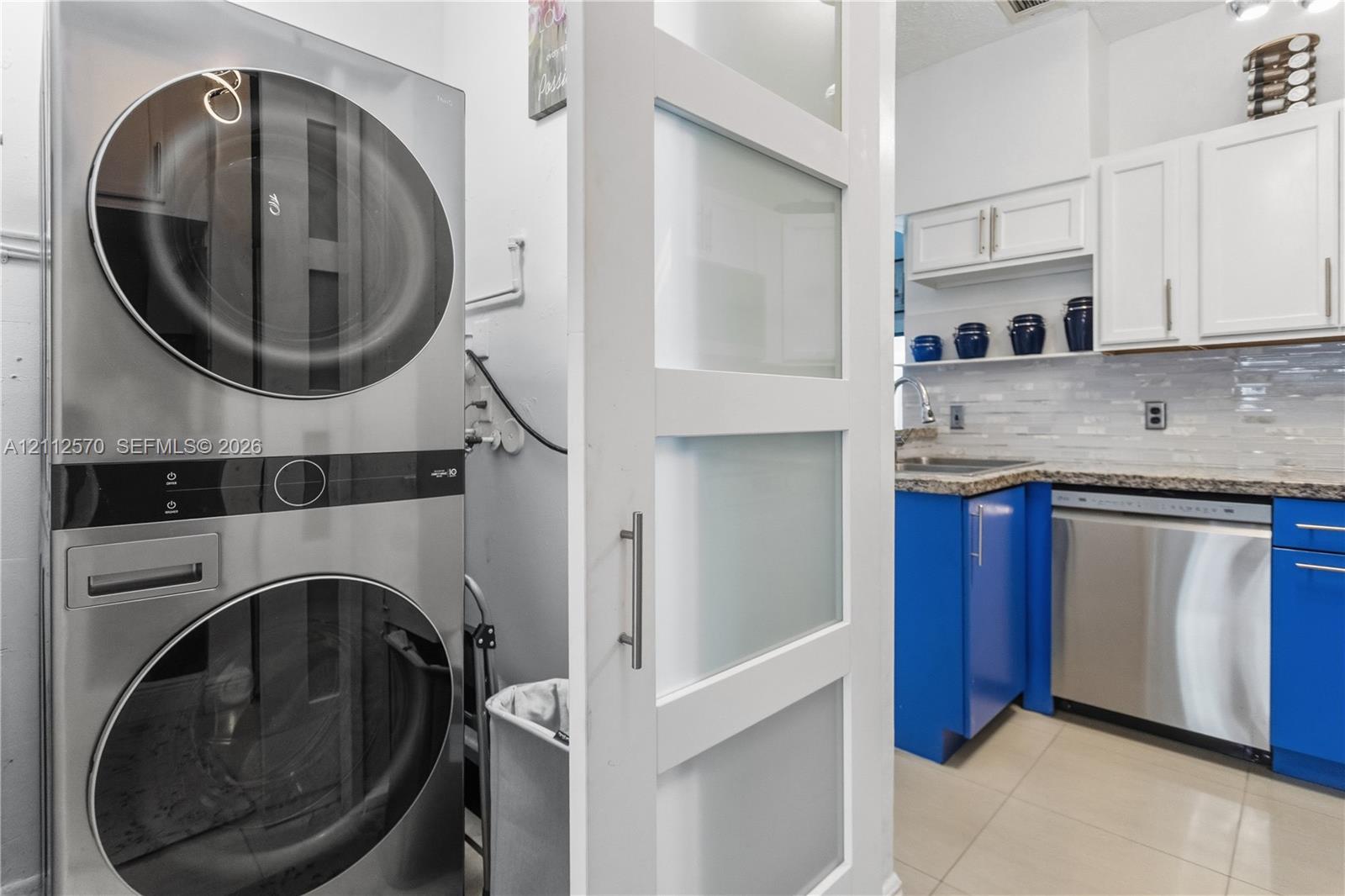 Laundry area featuring a stackable stainless steel appliance unit and a white sliding door with frosted glass panels, paired with a kitchen showcasing blue lower cabinetry, white upper cabinetry, tiled backsplash, and stainless steel dishwasher