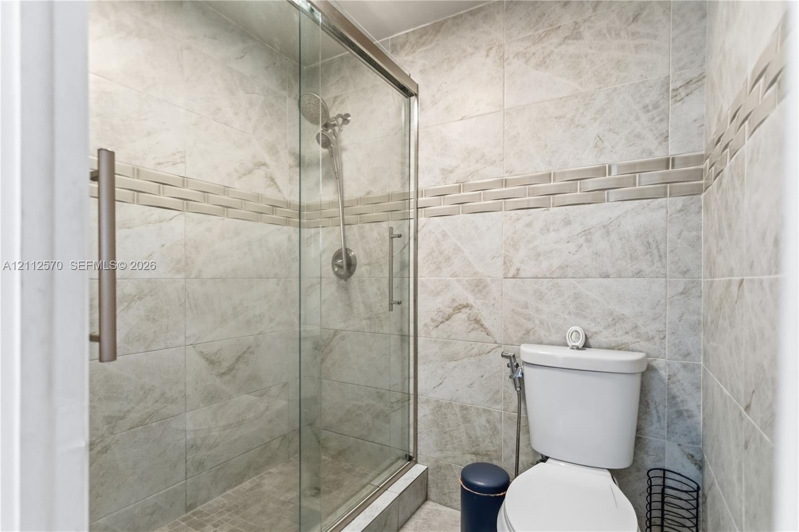 Walk-in shower with a glass enclosure, brushed nickel handheld showerhead, floor-to-ceiling stone-patterned tile, and decorative horizontal tile accent border
