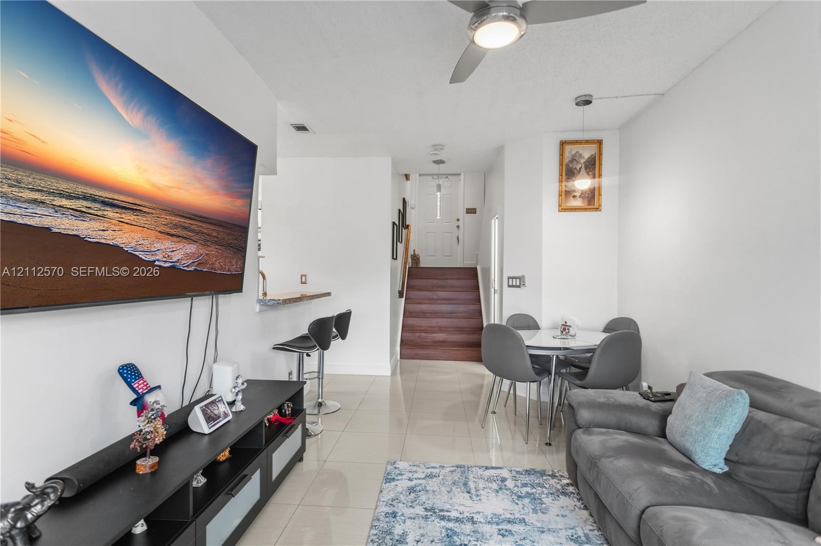 Open-concept interior featuring large-format tile flooring, a recessed breakfast bar, a ceiling fan with integrated lighting, and a staircase with wood-finish treads