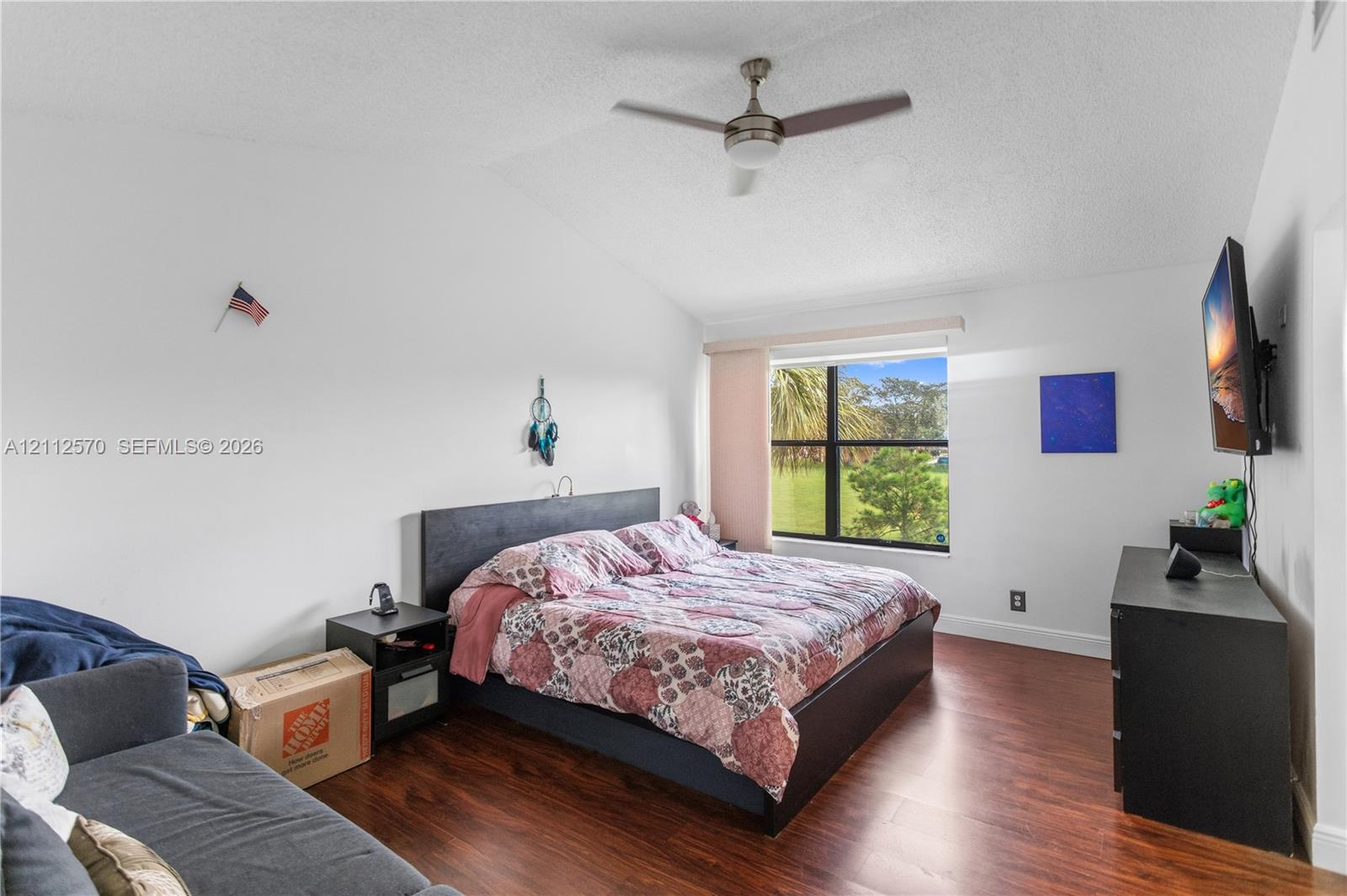 Vaulted ceiling with a central brushed-nickel ceiling fan, wood-finish flooring, and a large window providing natural light