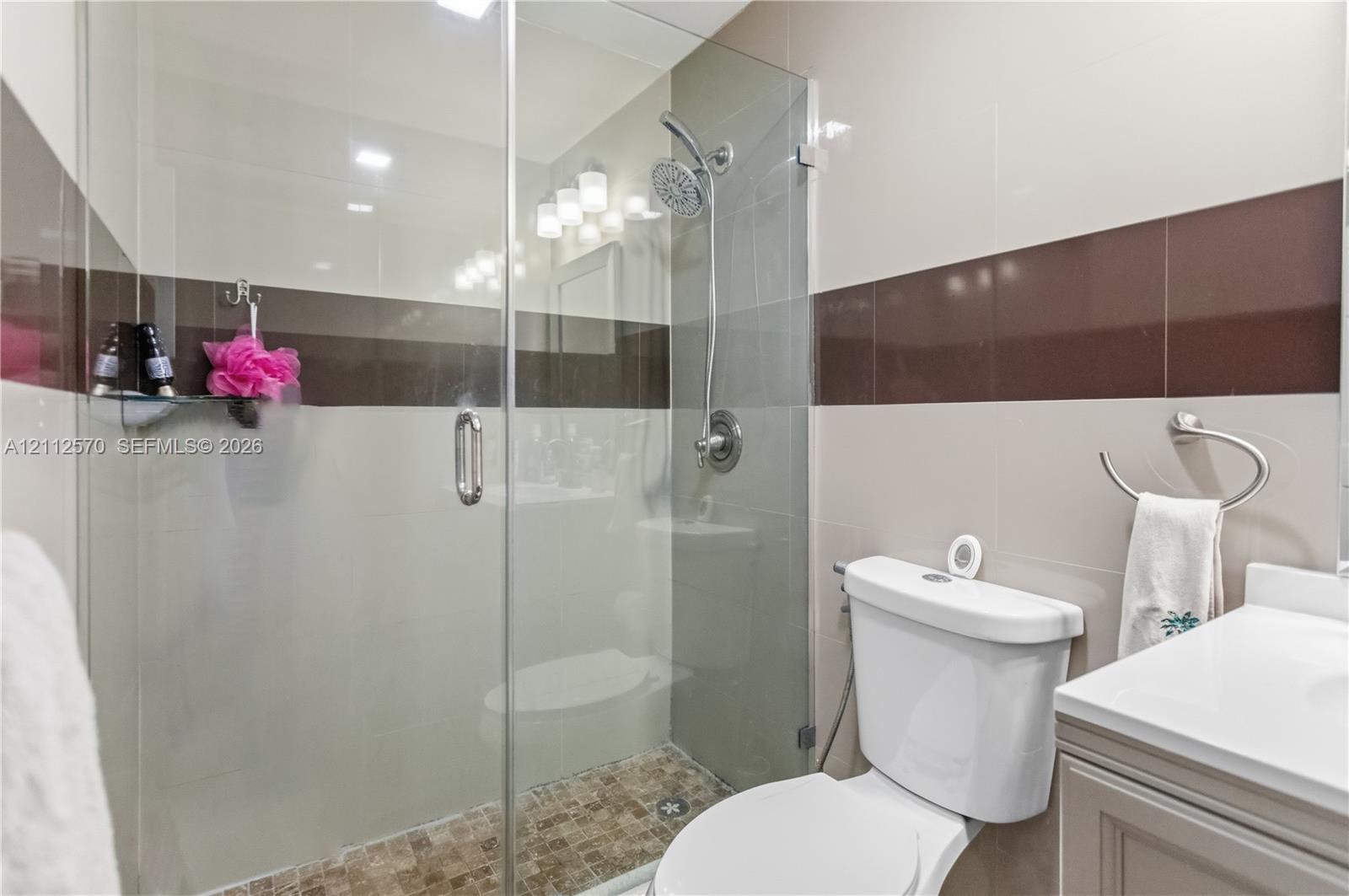 Walk-in shower featuring a glass enclosure, rainfall showerhead, and mosaic tile flooring, accented by dual-tone wall tiling and a neutral-toned vanity