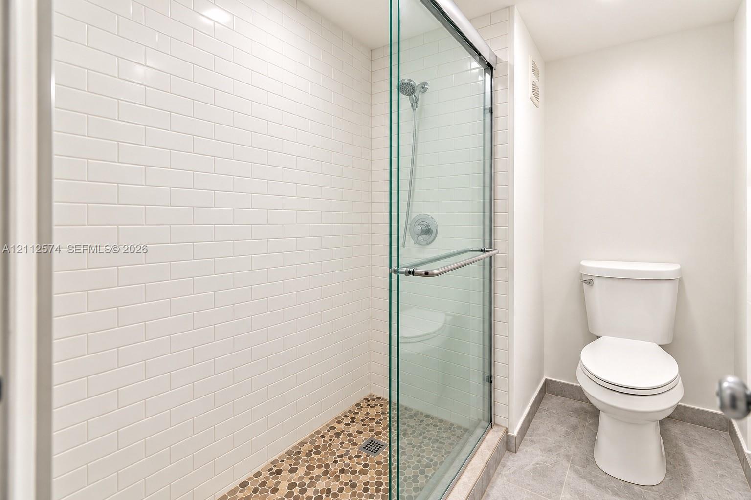 Walk-in shower featuring a frameless glass enclosure, white subway tile walls, and a pebble-stone floor, accented by a dual-function shower head and a standard toilet