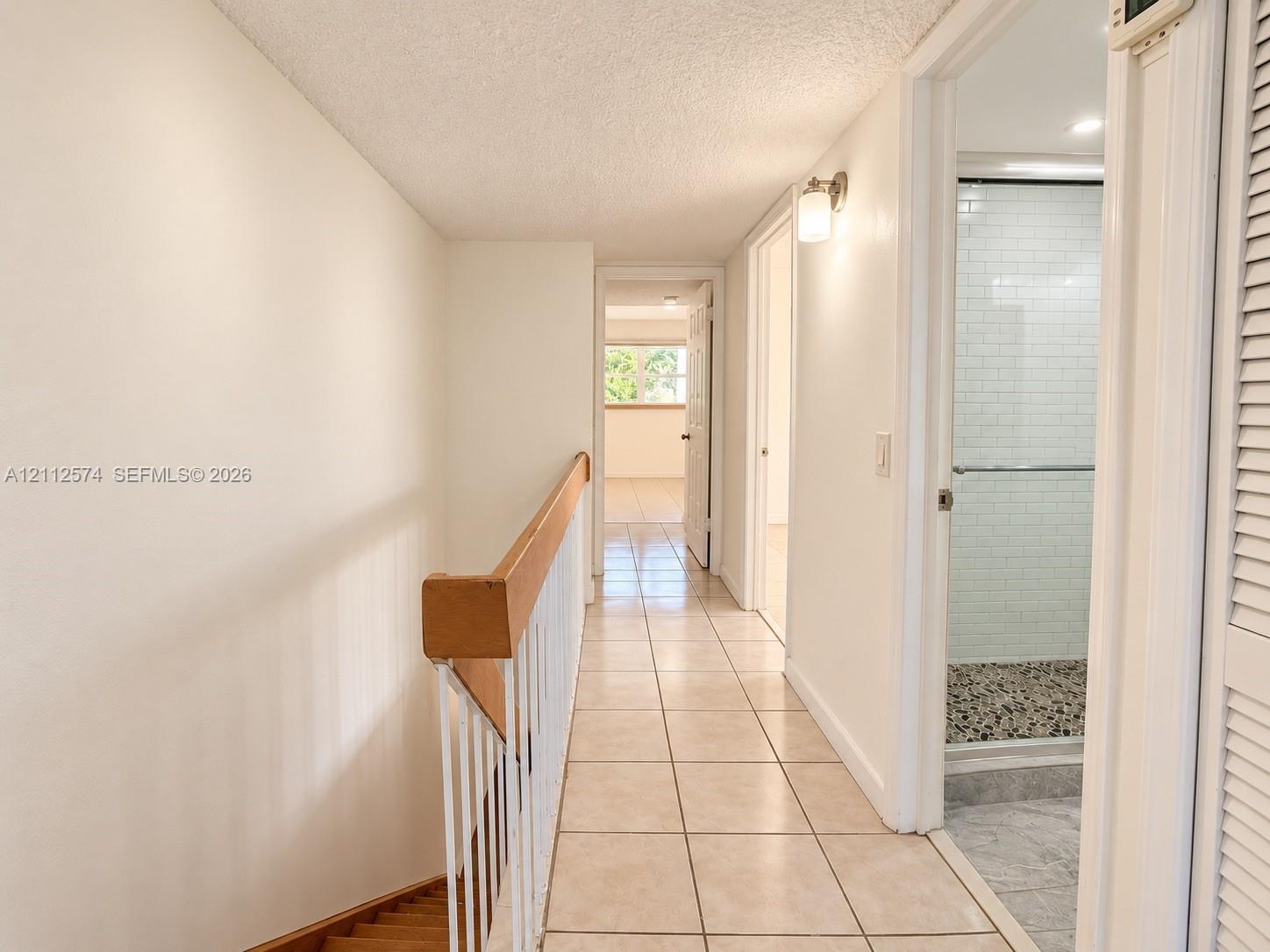 Hallway featuring large-format tile flooring, a wood-capped staircase railing with vertical white spindles, and a bathroom entry with white subway tile shower surround and stone-texture shower flooring