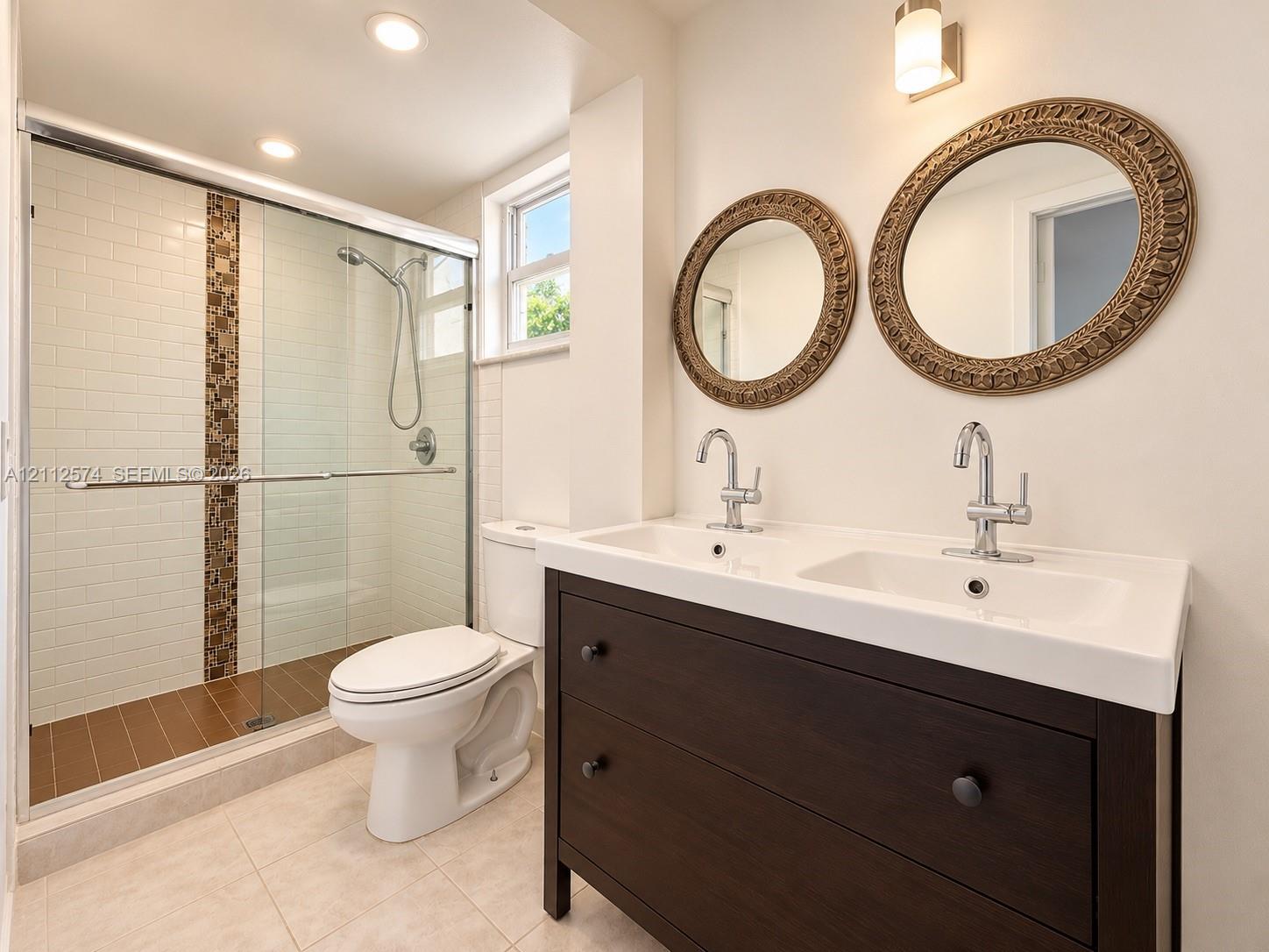 Double vanity with a wood-finish cabinet, rectangular sinks, and chrome faucets, featuring a glass-enclosed shower with white subway tile, a decorative vertical tile inlay, and a hand-held shower fixture