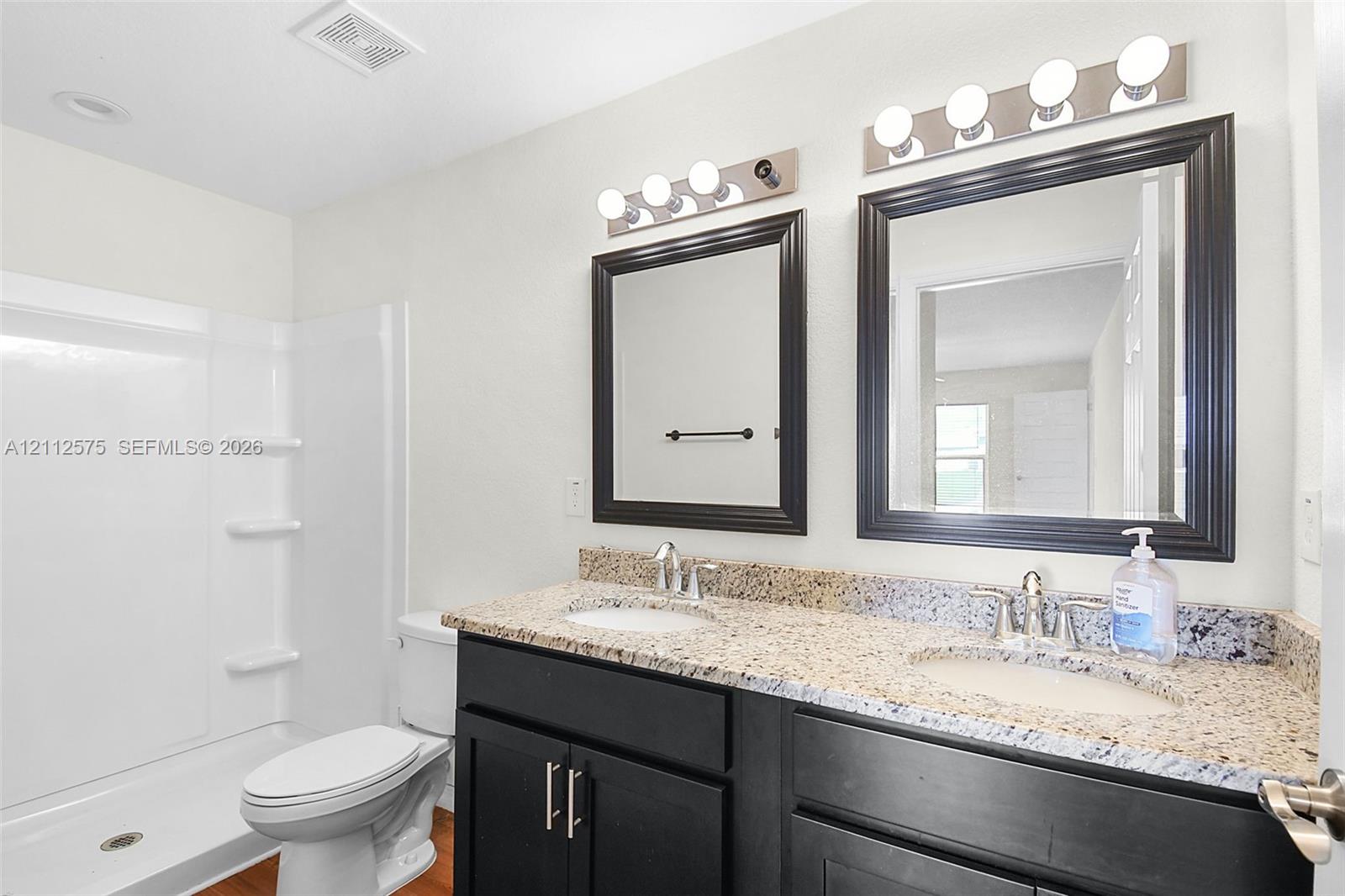 Dual-sink vanity with a stone-finish countertop, dark cabinetry, matching wall-mounted mirrors, and dual four-light fixtures, accompanied by a fiberglass shower surround