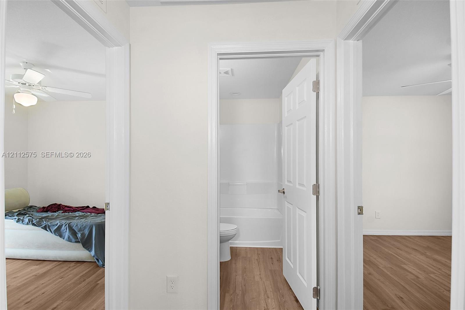 Interior hallway featuring wood-finish flooring, white six-panel doors, and a bathroom equipped with a white tub-shower surround