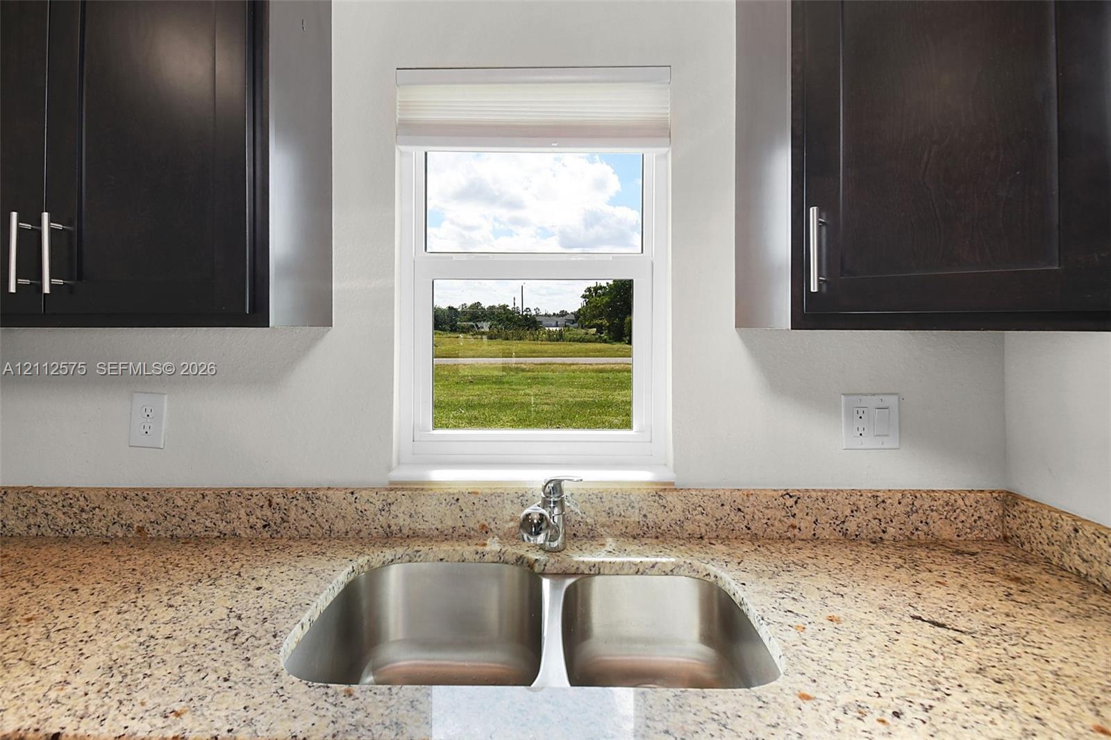 Deep-toned cabinetry featuring vertical pulls, speckled stone countertops, dual-basin stainless steel sink, and a centralized window with integrated shade