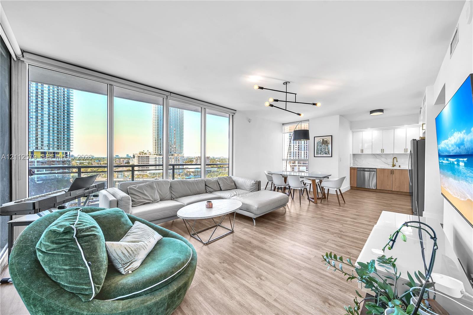 Floor-to-ceiling windows, wood-finish flooring, linear chandelier, minimalist kitchen cabinetry, and stone-finish backsplash Floor-to-ceiling windows, wood-finish flooring, linear chandelier, minimalist kitchen cabinetry, and stone-finish backsplash