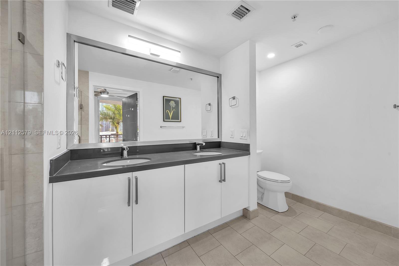 Double vanity with a dark solid-surface countertop, dual undermount sinks, sleek white cabinetry, and a large wall-to-wall mirror with integrated lighting