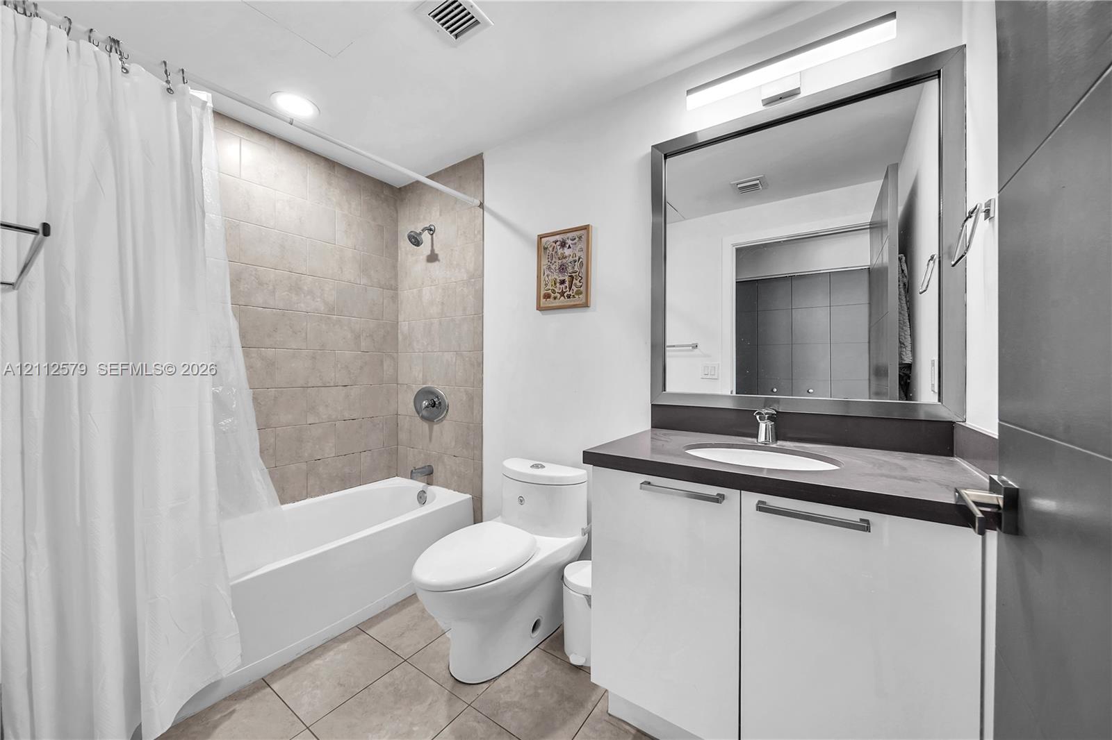 Bathroom featuring a high-gloss white vanity with a dark solid-surface countertop, rectangular mirror with a metal frame, tile-surround bathtub, and large-format floor tiling