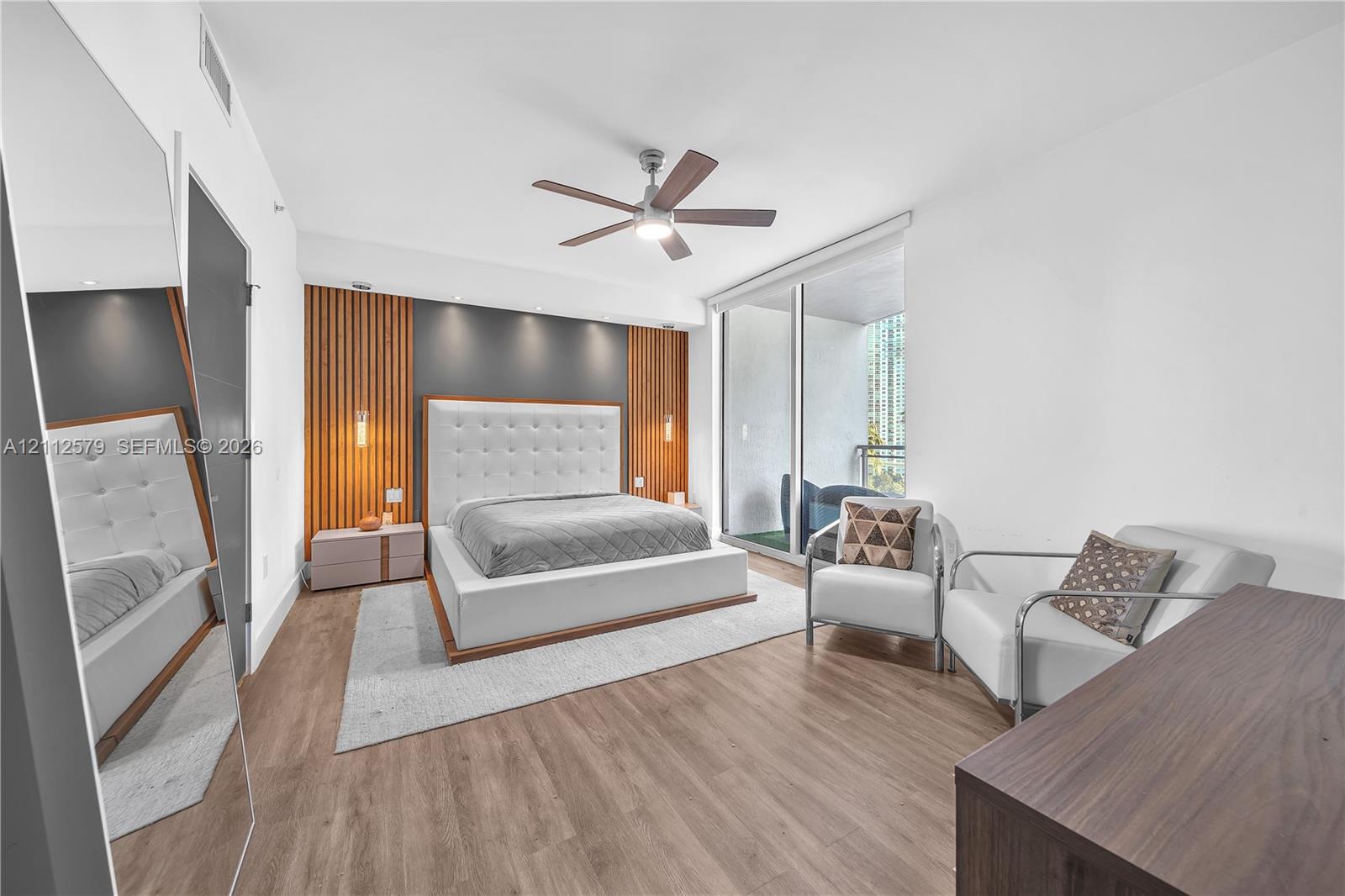 Wood-finish flooring, vertical slat accent wall with recessed lighting, ceiling fan, and full-height sliding glass doors opening to a balcony Wood-finish flooring, vertical slat accent wall with recessed lighting, ceiling fan, and full-height sliding glass doors opening to a balcony
