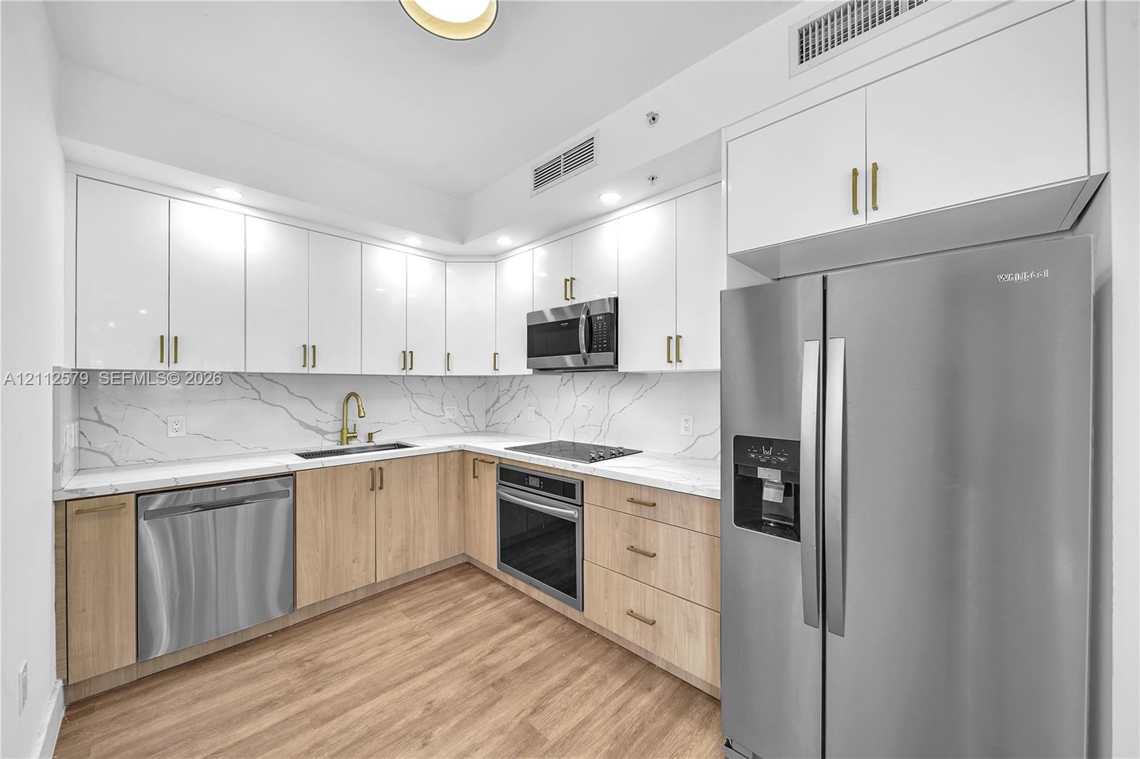 Kitchen featuring high-gloss white upper cabinetry, wood-finish base cabinetry, veined stone-look backsplash and countertops, brushed brass hardware, and stainless steel appliances Kitchen featuring high-gloss white upper cabinetry, wood-finish base cabinetry, veined stone-look backsplash and countertops, brushed brass hardware, and stainless steel appliances