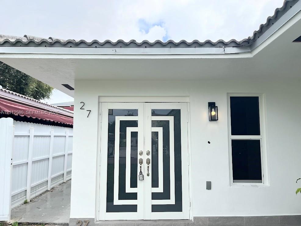 White stucco exterior featuring a double-door entry with geometric detailing, tile roof, vertical single-hung window, and wall-mounted exterior lighting White stucco exterior featuring a double-door entry with geometric detailing, tile roof, vertical single-hung window, and wall-mounted exterior lighting