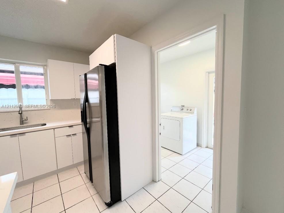 Kitchen featuring white cabinetry, light-toned countertops, a stainless steel refrigerator, and tiled flooring extending into an additional room containing laundry hookups Kitchen featuring white cabinetry, light-toned countertops, a stainless steel refrigerator, and tiled flooring extending into an additional room containing laundry hookups