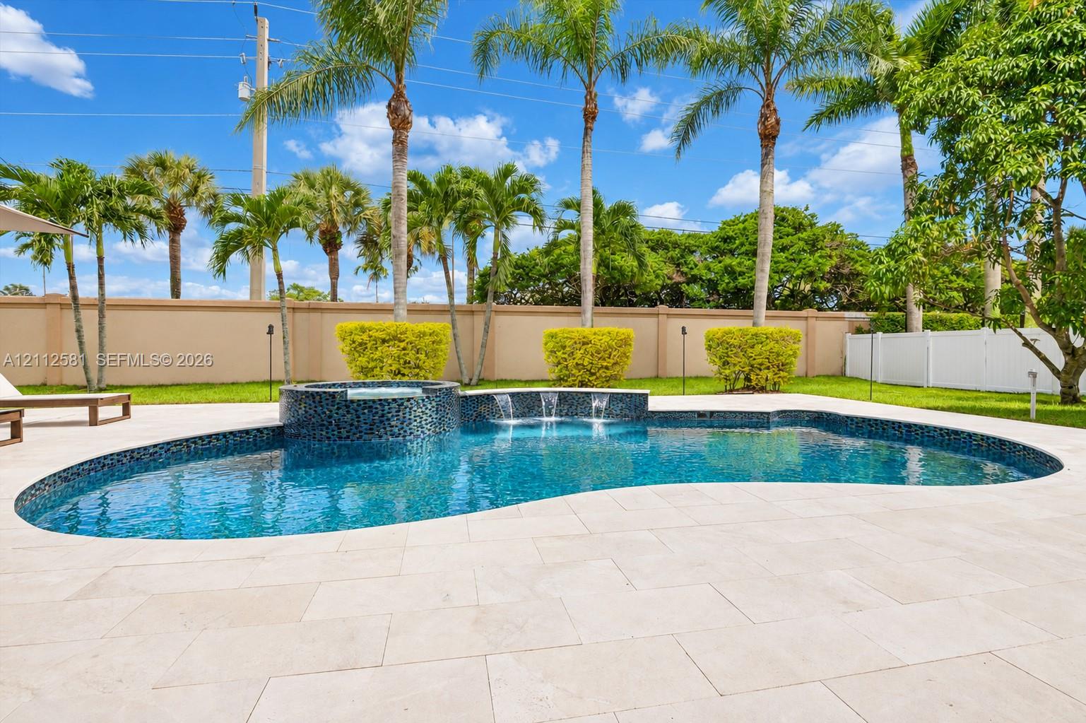 Freeform swimming pool with a raised spillover spa, water features, tiled pool finish, and expansive light-toned paver deck, all set within a privacy-walled yard featuring mature palm trees and manicured shrubbery