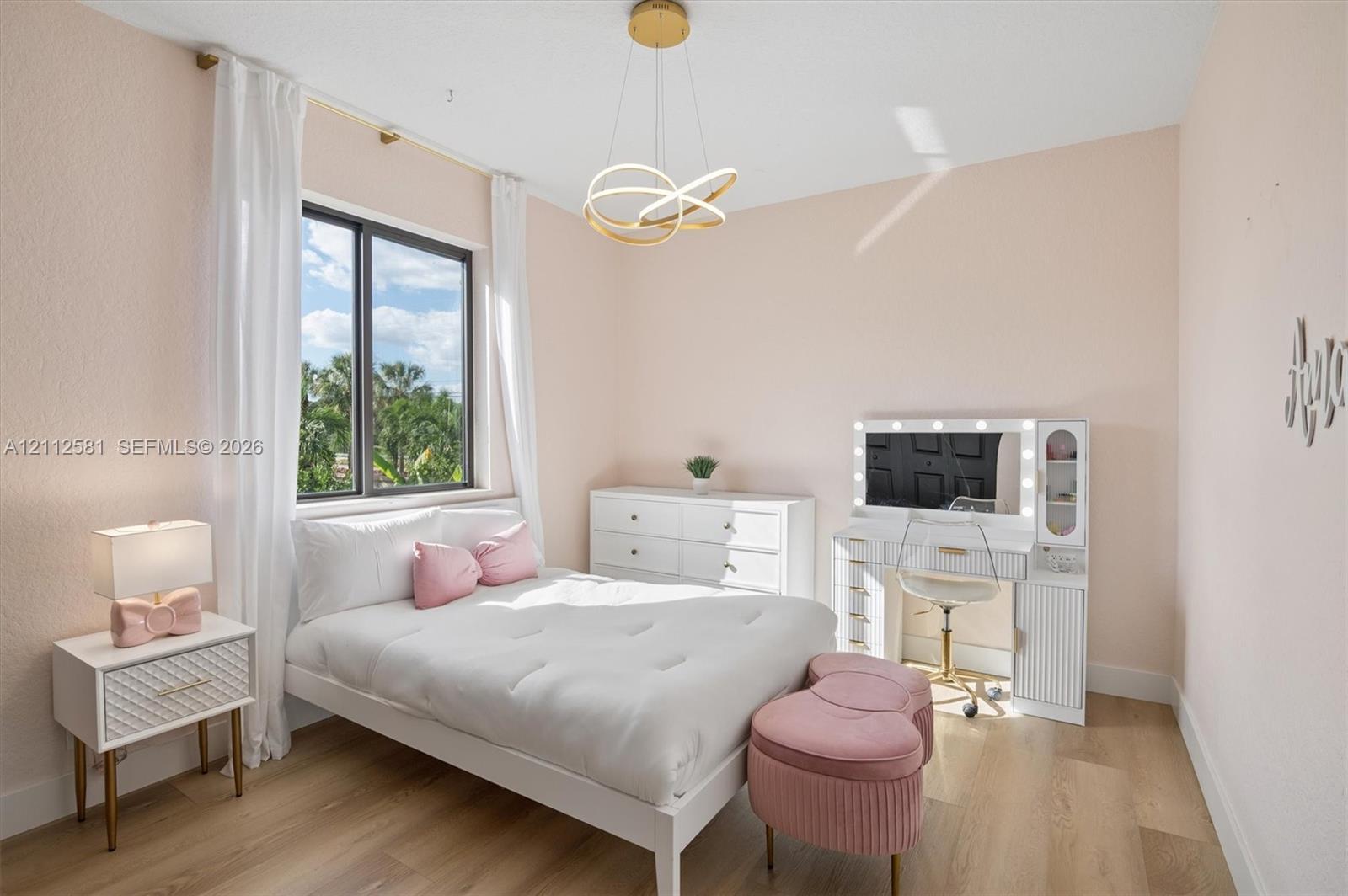 Bedroom featuring wood-finish flooring, a large window with exterior tropical scenery, and a modern gold-finish geometric pendant light