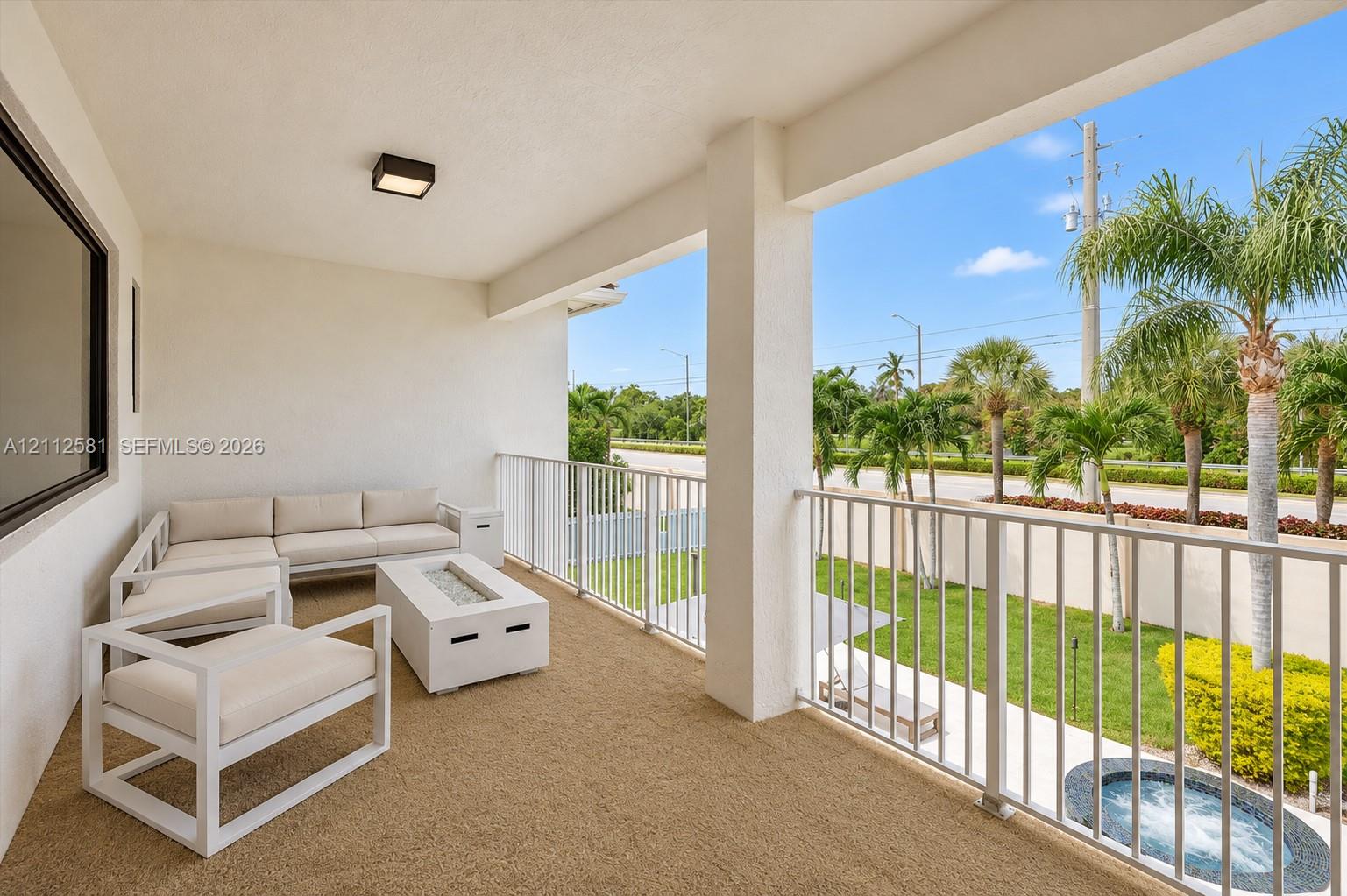 Covered balcony featuring textured flooring, white metal railing, flush-mount lighting, and exterior access to a fenced rear yard with a built-in spa
