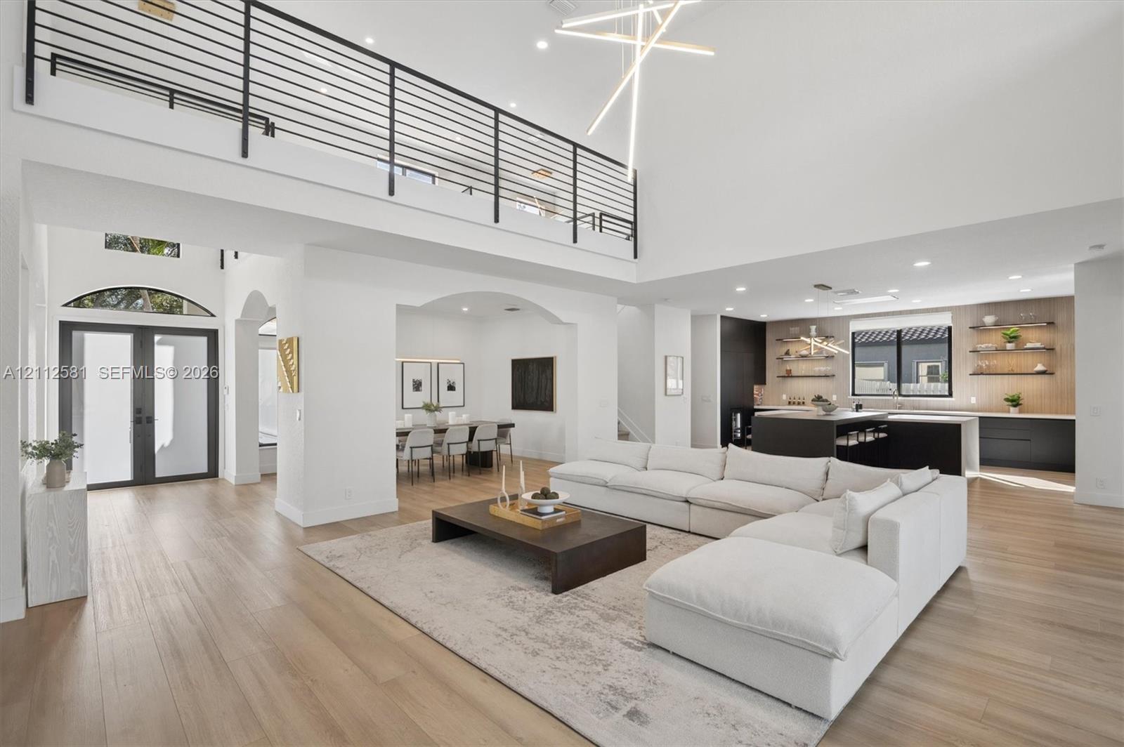 Double-height ceiling with geometric pendant lighting, wood-finish flooring, metal horizontal-bar mezzanine railing, and kitchen featuring dark-tone cabinetry with open wood shelving