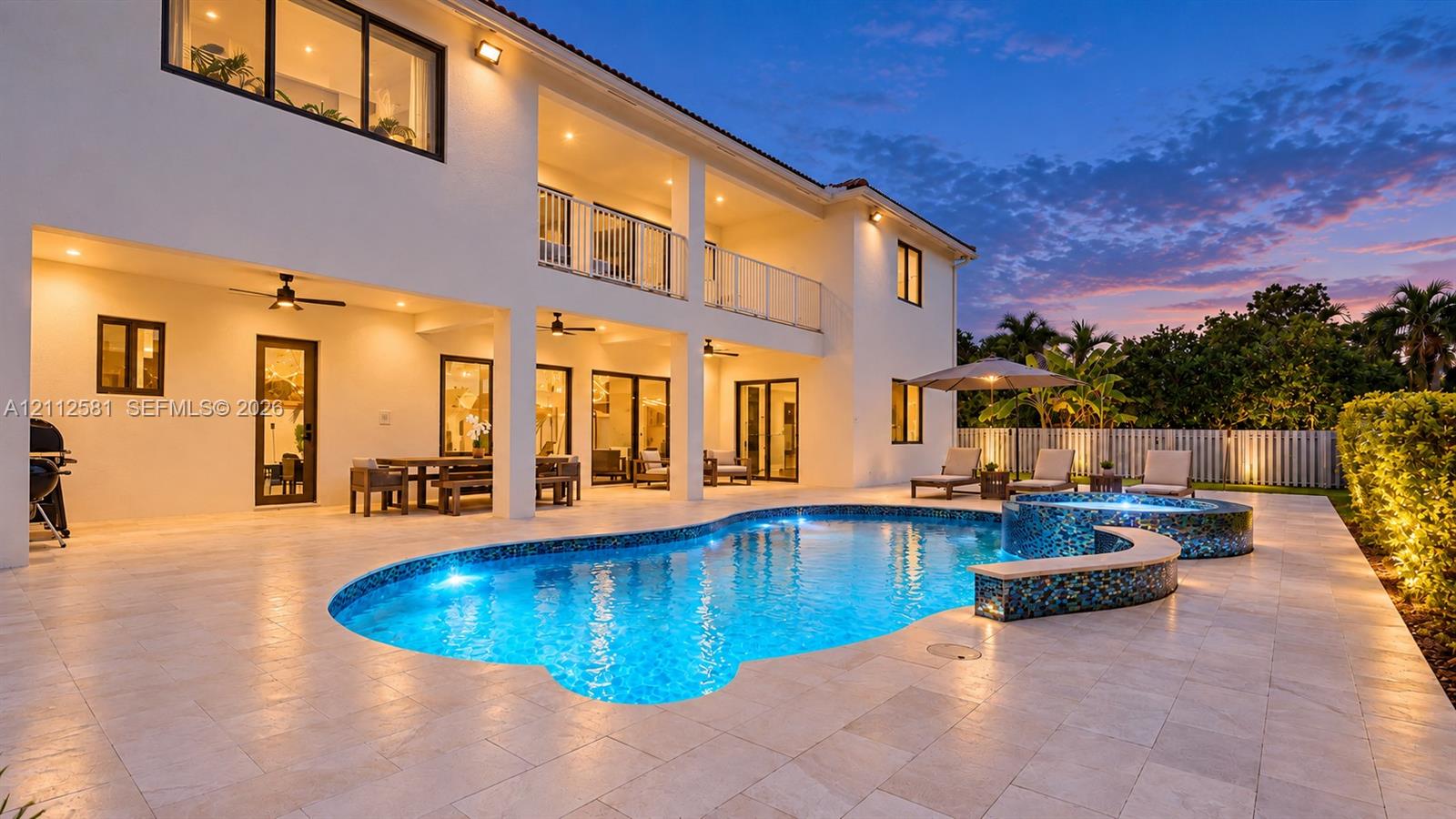 Two-story exterior featuring a stone-paved patio, a custom-shaped pool with integrated spa, multiple ceiling fans, and a second-level balcony with metal railings