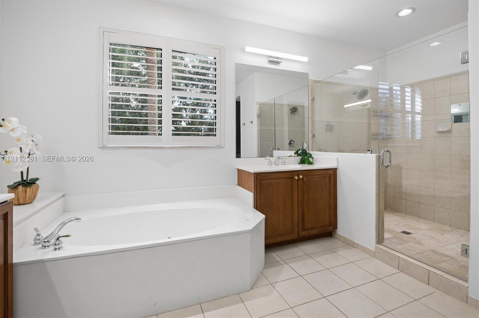 Built-in soaking tub with a deck-mounted faucet, wood cabinetry vanity, large wall mirror, glass-enclosed shower with tile surround, and plantation shutters