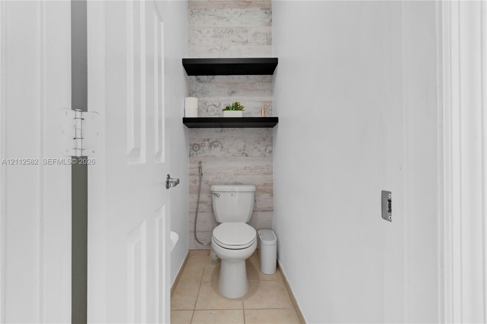 Restroom featuring ceramic tile flooring, two black floating shelves, decorative wall surfacing, and a wall-mounted bidet sprayer