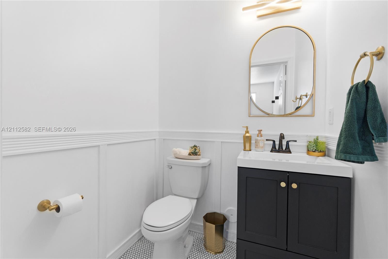Powder room featuring wainscoting, a dark vanity cabinet with a stone-top sink, arched gold-rimmed mirror, patterned floor tile, and gold hardware fixtures