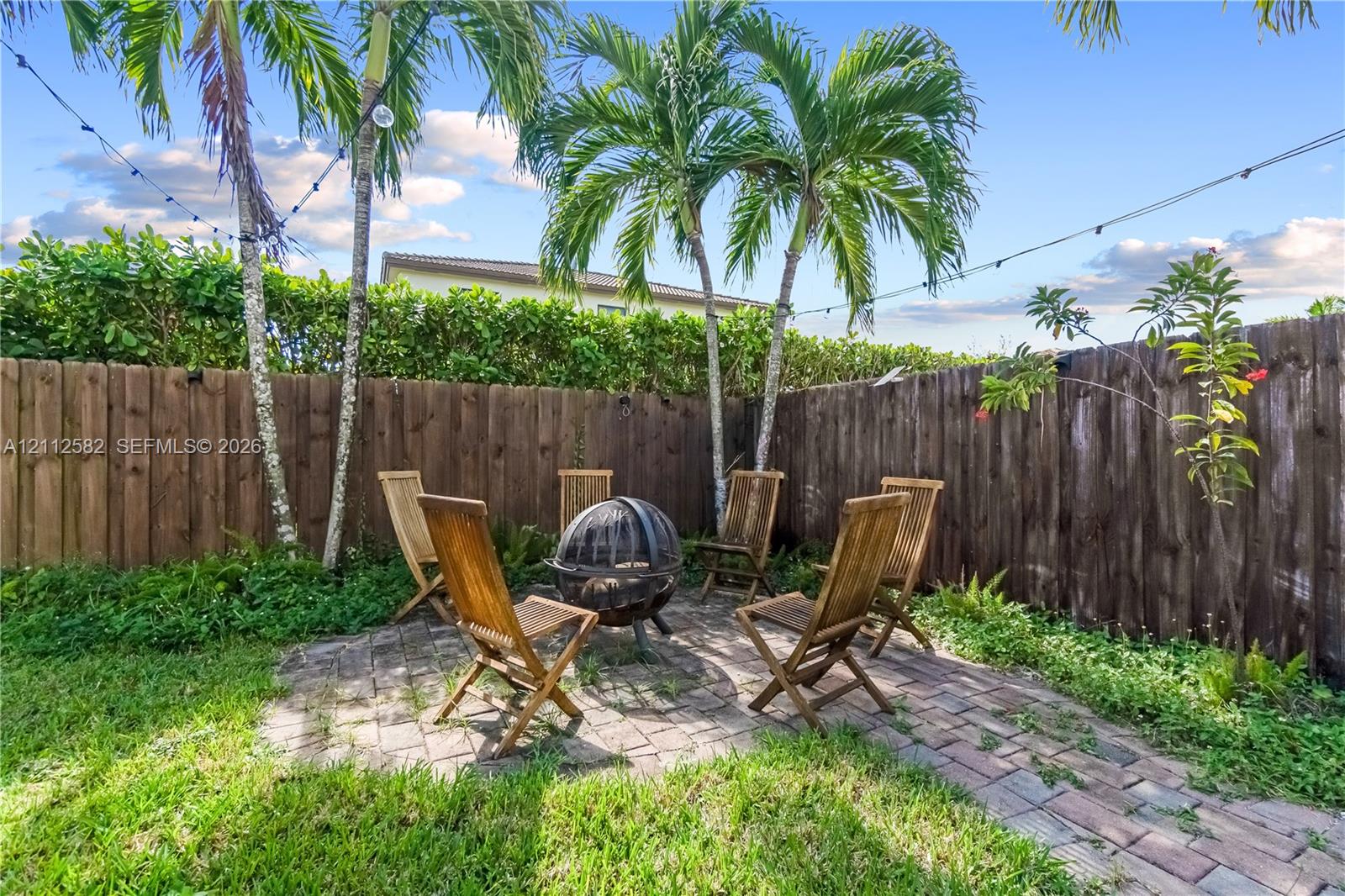 Backyard featuring a brick paver patio, wood privacy fencing, mature palm trees, and overhead string lighting