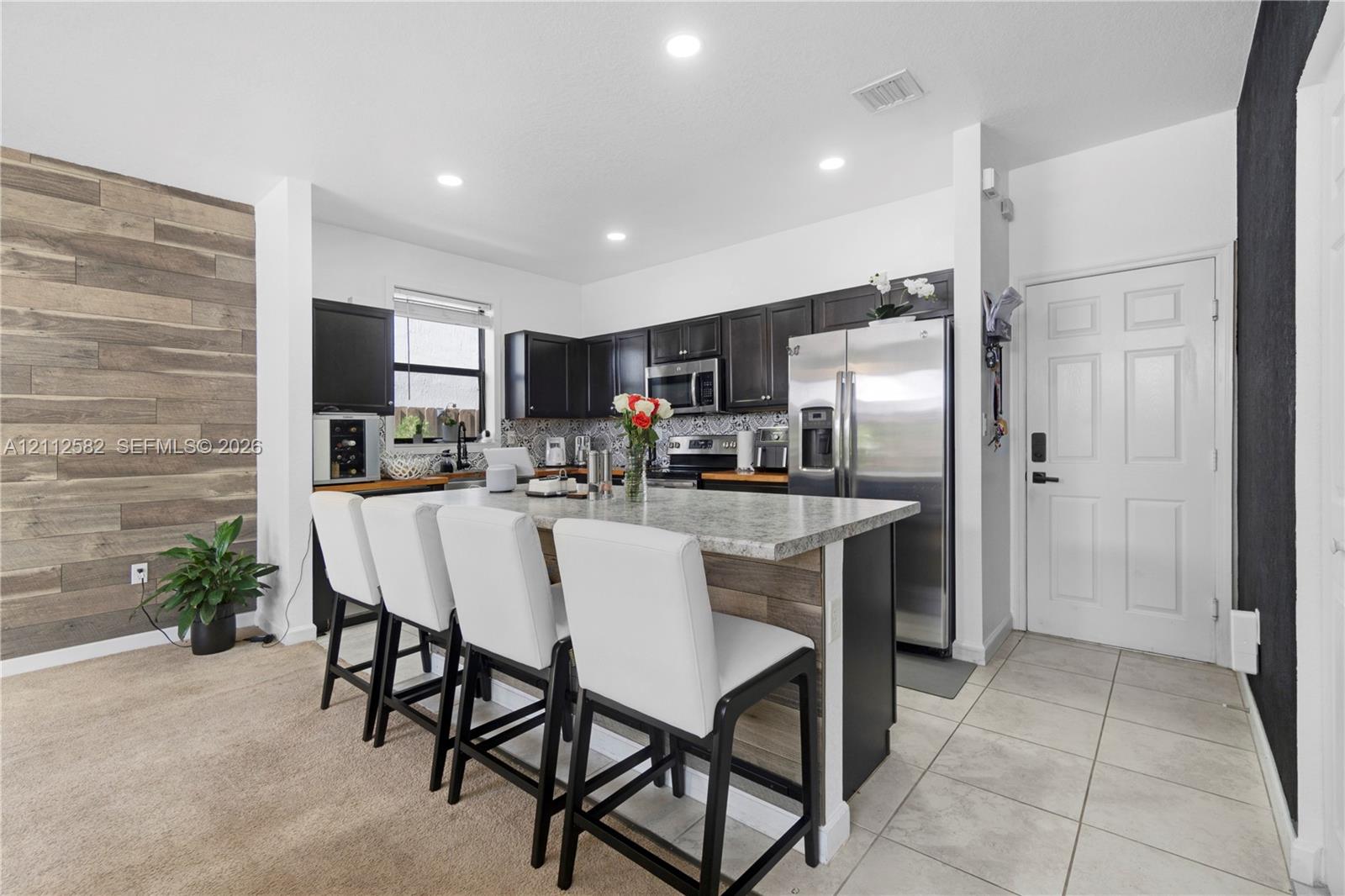 Kitchen featuring a stone-composite countertop island, dark cabinetry, recessed lighting, and a wood-finish accent wall Kitchen featuring a stone-composite countertop island, dark cabinetry, recessed lighting, and a wood-finish accent wall