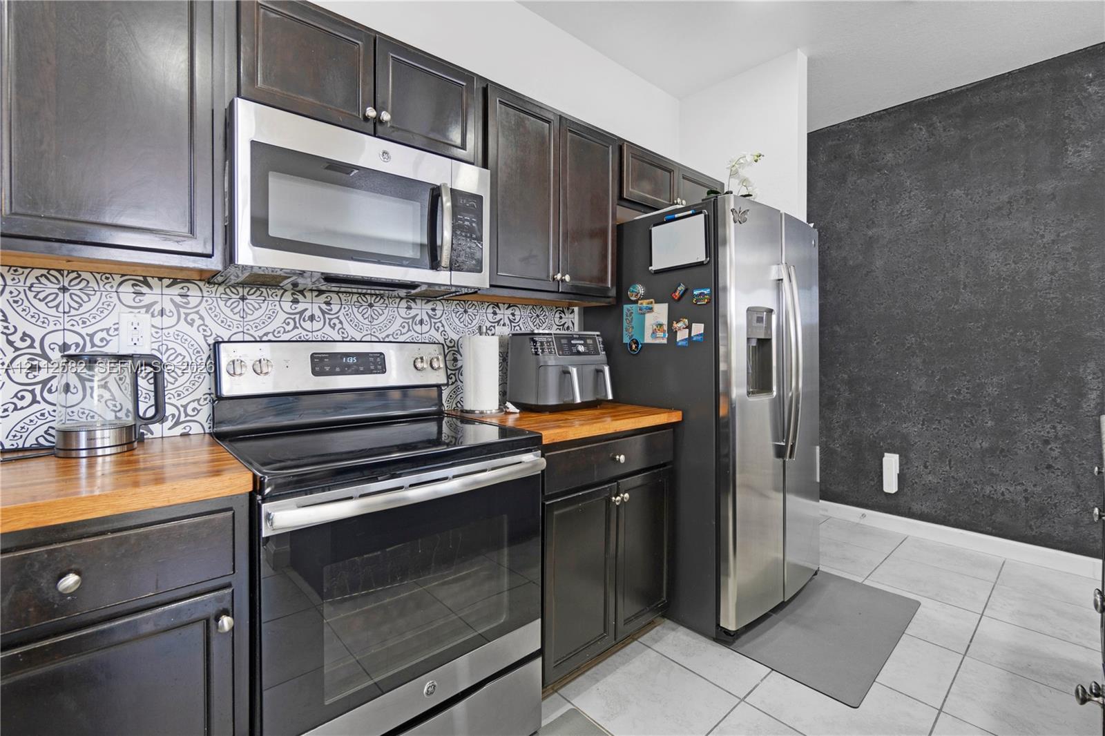 Kitchen featuring dark-finish cabinetry, butcher-block countertops, stainless steel appliances, decorative patterned backsplash, and tile flooring Kitchen featuring dark-finish cabinetry, butcher-block countertops, stainless steel appliances, decorative patterned backsplash, and tile flooring
