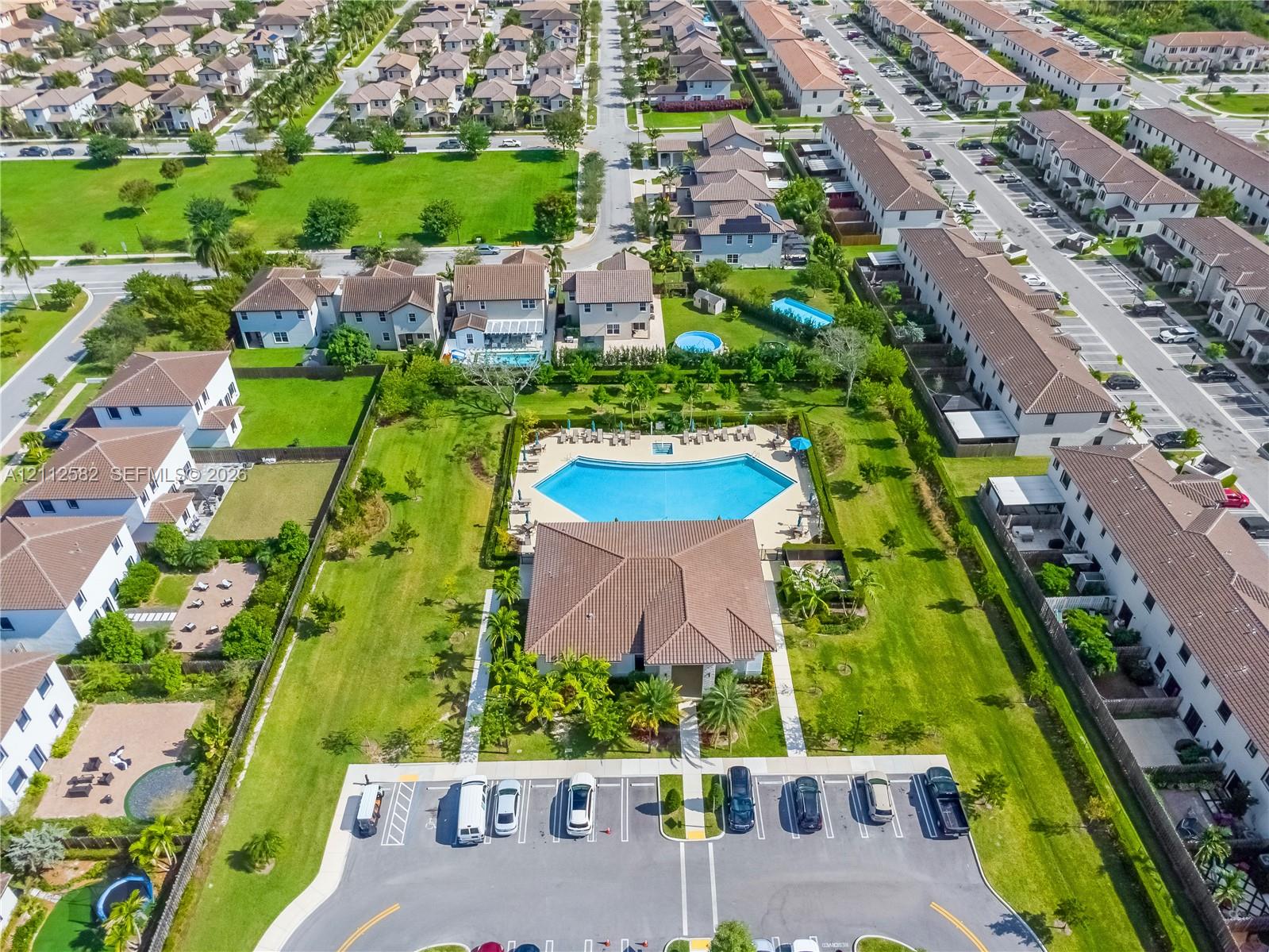Aerial perspective of a community recreational center featuring a swimming pool, expansive green space, mature landscaping, and dedicated surface parking