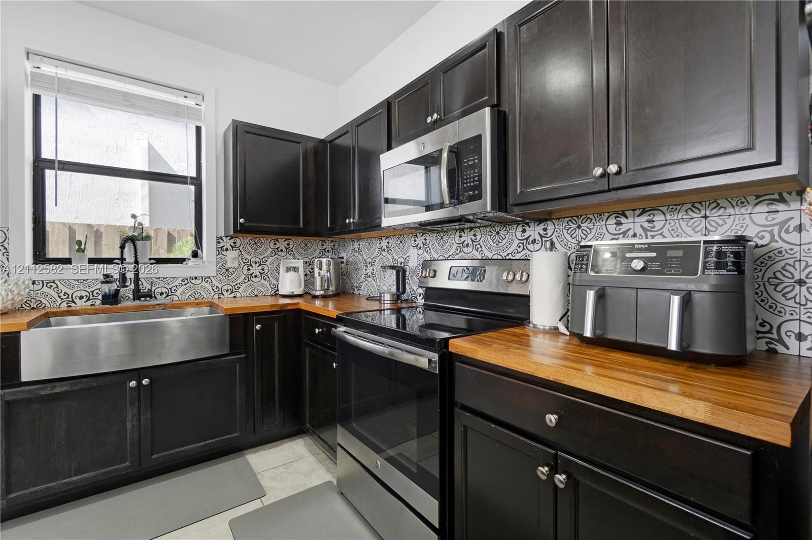 Kitchen featuring dark cabinetry, butcher block countertops, stainless steel farmhouse sink with a gooseneck faucet, stainless steel appliances, and patterned tile backsplash Kitchen featuring dark cabinetry, butcher block countertops, stainless steel farmhouse sink with a gooseneck faucet, stainless steel appliances, and patterned tile backsplash