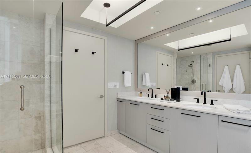 Dual-sink vanity with light-gray cabinetry, stone countertops, matte black fixtures, full-width mirror, and walk-in tiled shower with a glass enclosure