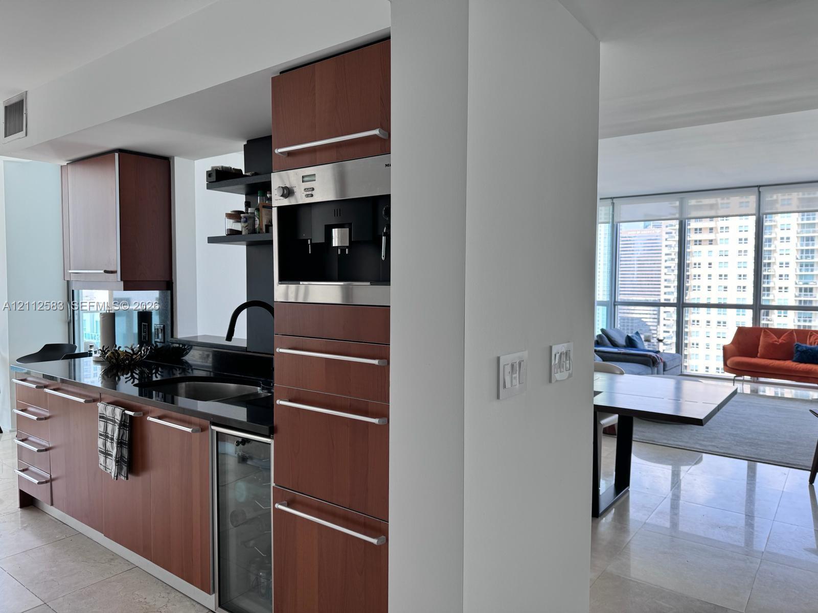 Kitchen featuring wood-veneer cabinetry, integrated coffee station, dark stone countertops, under-counter wine refrigerator, and polished stone flooring