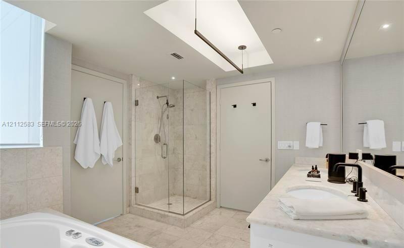 Bathroom featuring a skylight, walk-in glass-enclosed shower with stone tile surround, dual-sink vanity with light stone countertop, and deep soaking tub