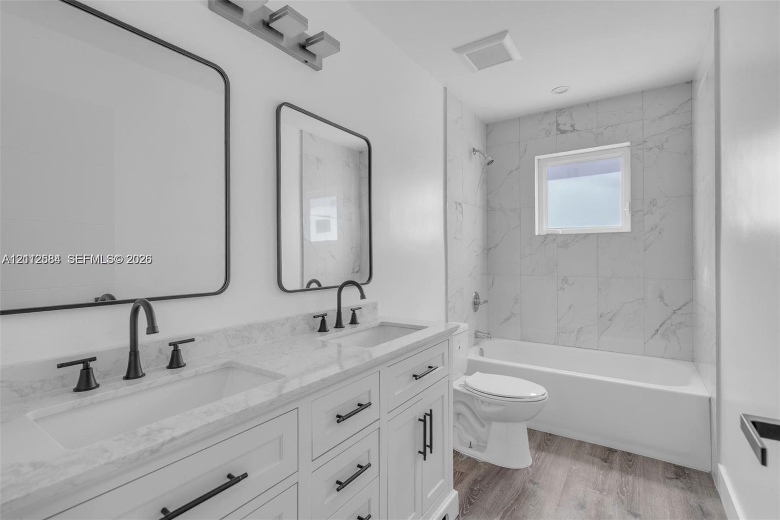 Dual-sink vanity with stone-finish countertop, white shaker-style cabinetry, matte black hardware, dual rectangular mirrors, and a tub-shower combination featuring floor-to-ceiling tile surround
