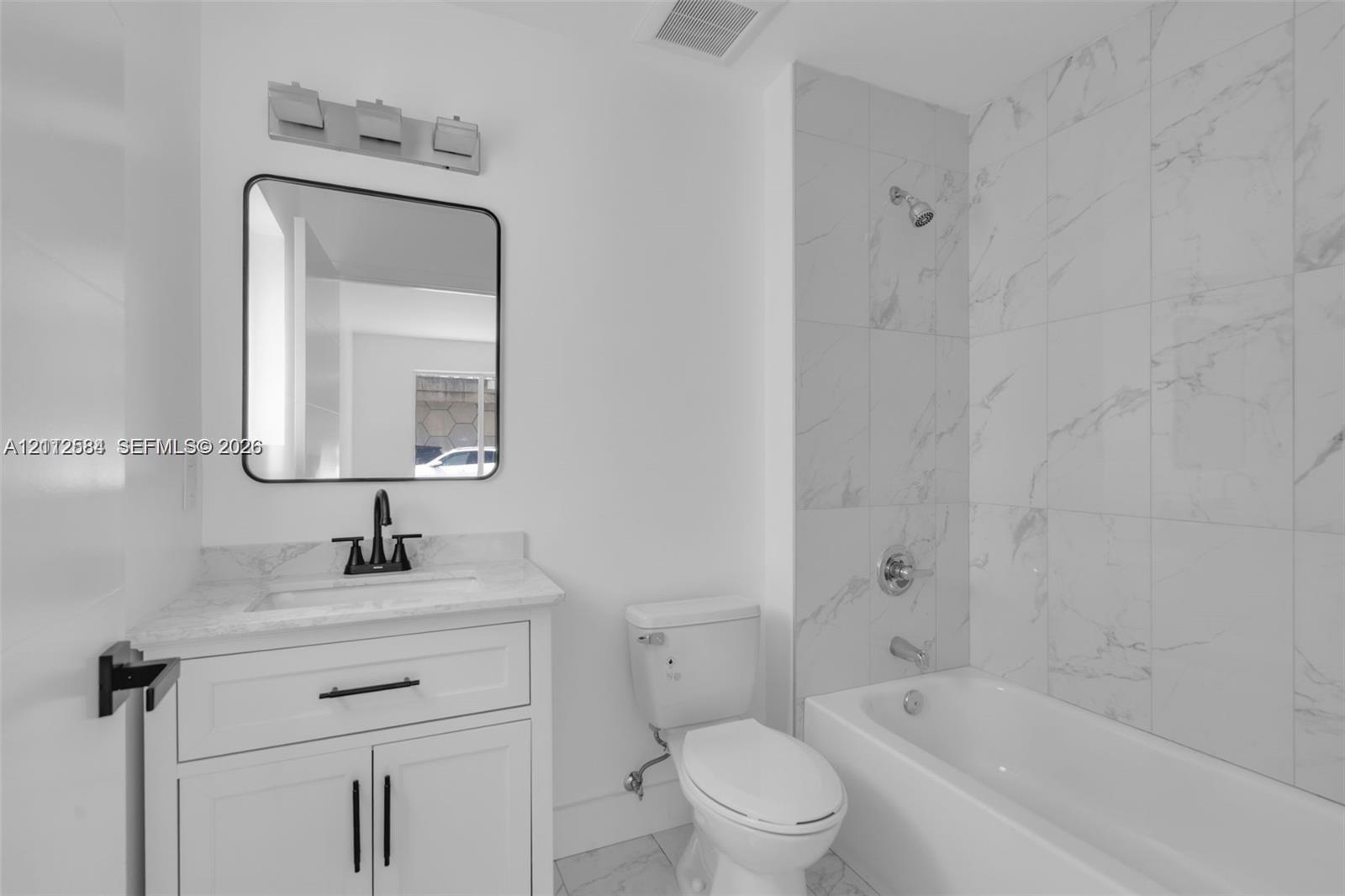 Bathroom featuring white shaker-style cabinetry, stone-surface countertop, matte black fixtures, and floor-to-ceiling tile surrounding the tub