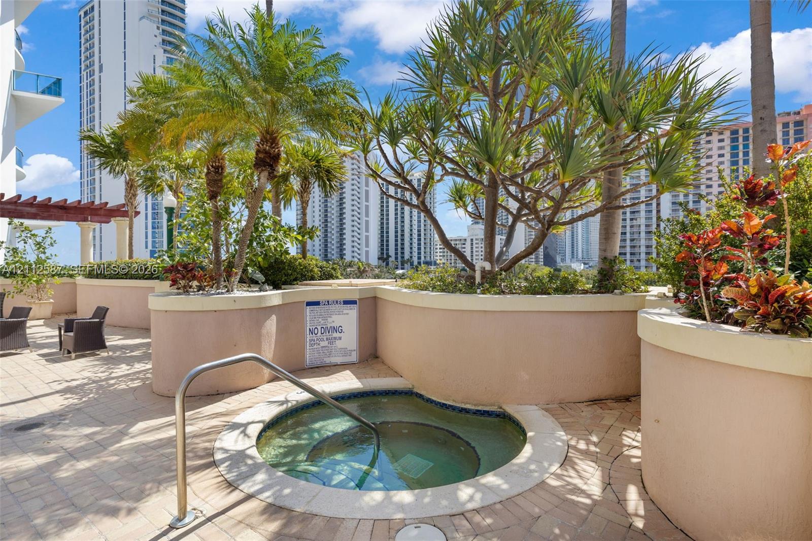 In-ground spa with a metal handrail, paver-stone decking, raised concrete planters, and mature tropical landscaping