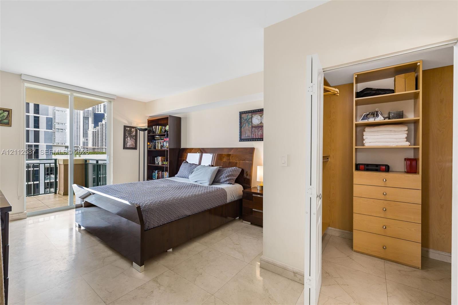 Sliding glass doors leading to a private balcony, large-format tile flooring, and a walk-in closet featuring built-in wood-finish shelving and drawer storage