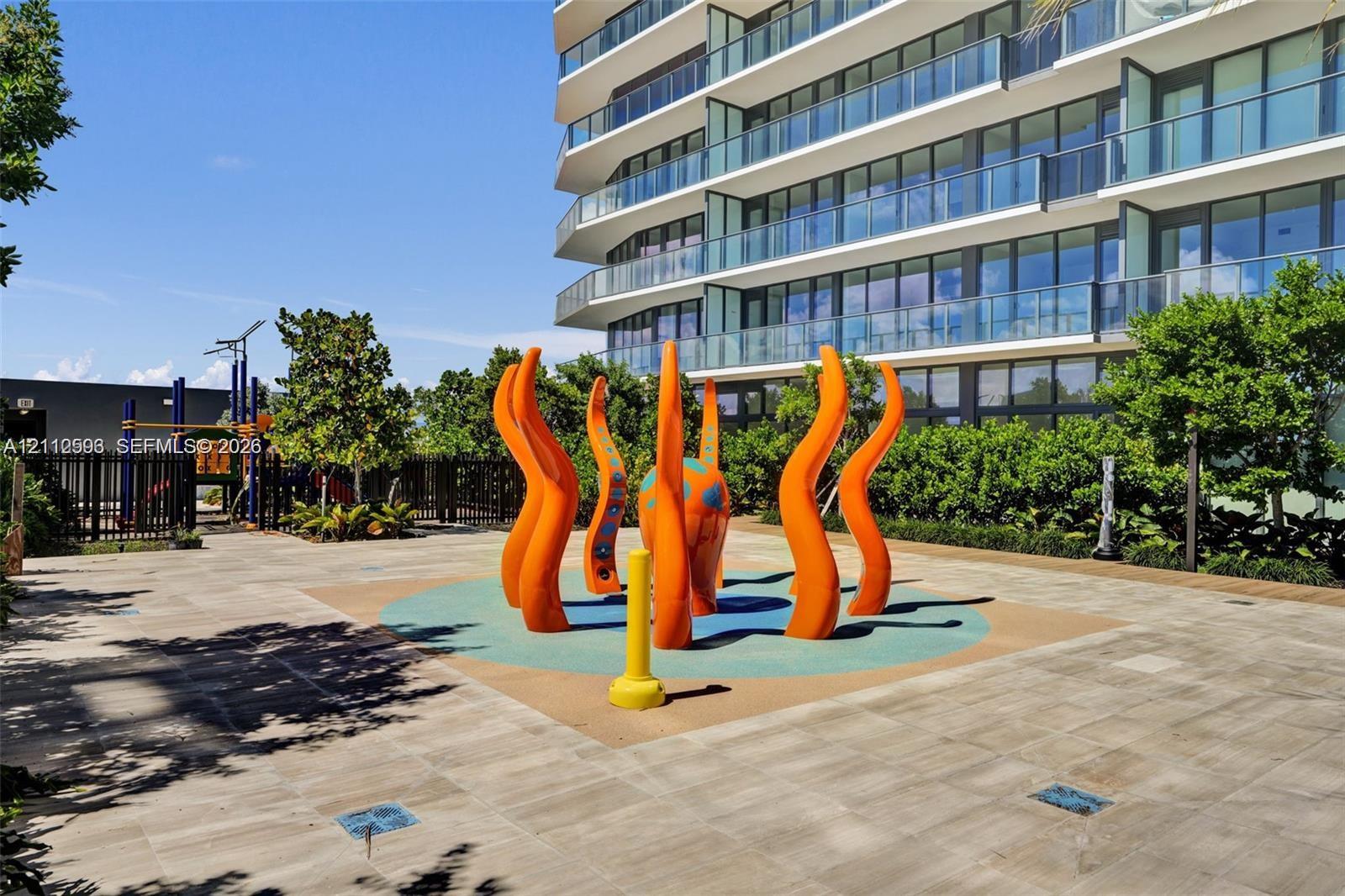 Outdoor recreation area featuring a paved terrace, colorful sculptural water feature, integrated rubberized ground surface, and perimeter landscaping