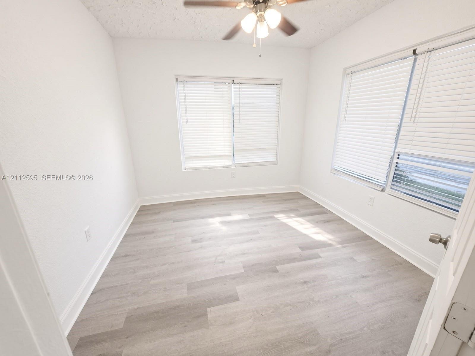 Room featuring wood-finish flooring, white walls, dual windows with blinds, and a ceiling fan with light fixture