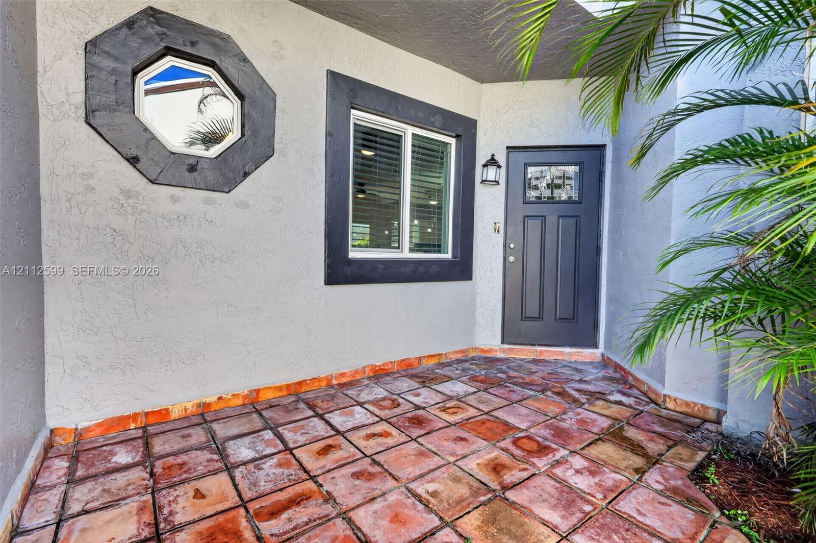 Covered entry featuring terra-cotta tile flooring, a solid-core front door with decorative glass inlay, an octagonal accent window, and dark-finished exterior trim