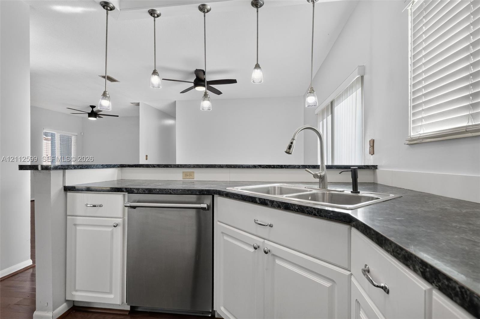 Kitchen featuring white cabinetry, dark stone-finish countertops, stainless steel dishwasher, dual-basin sink with pull-down faucet, and four glass pendant lights