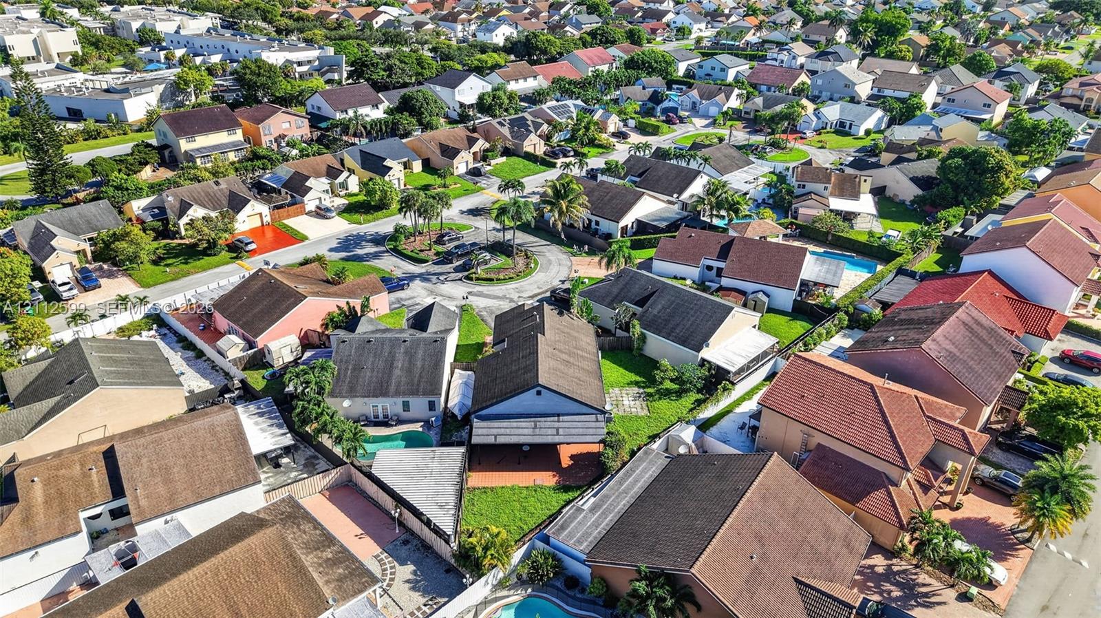 Aerial perspective of a residential neighborhood featuring multiple low-rise dwellings with tile roofing and paved driveways, surrounded by mature palm trees and manicured lawns