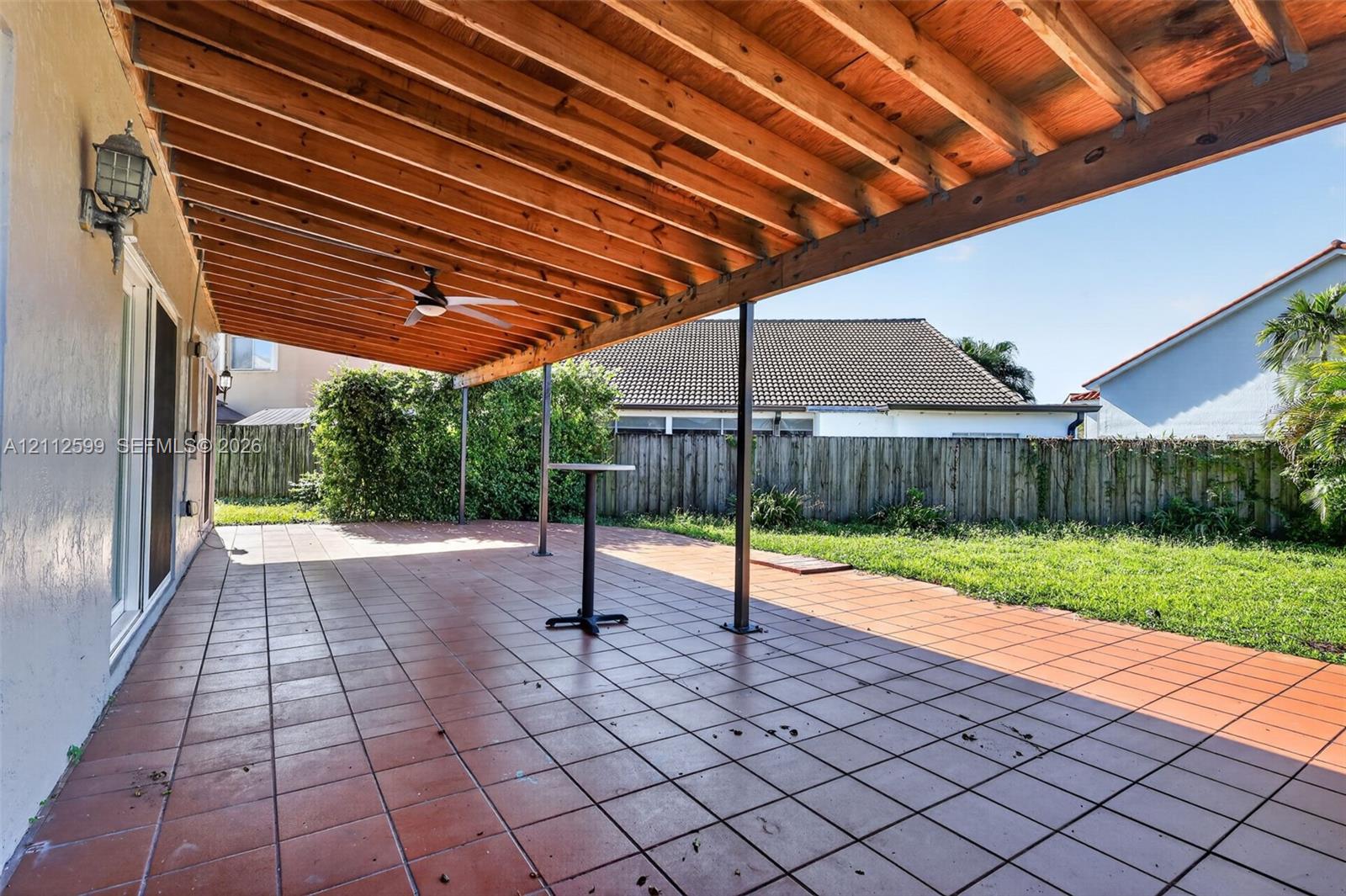 Covered rear patio featuring a wood-beam pergola, tiled flooring, integrated ceiling fan, and exterior wall-mounted lighting