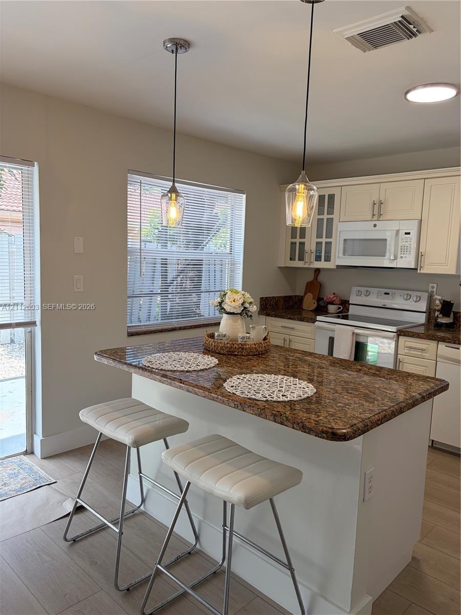 Kitchen featuring a center island with a granite countertop, off-white cabinetry, glass-front upper cabinet doors, and dual glass pendant light fixtures Kitchen featuring a center island with a granite countertop, off-white cabinetry, glass-front upper cabinet doors, and dual glass pendant light fixtures