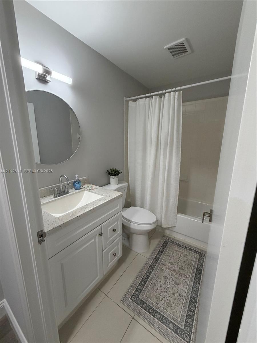 White cabinetry vanity with a speckled countertop, integrated basin, brushed nickel faucet, round wall-mounted mirror, and tile flooring
