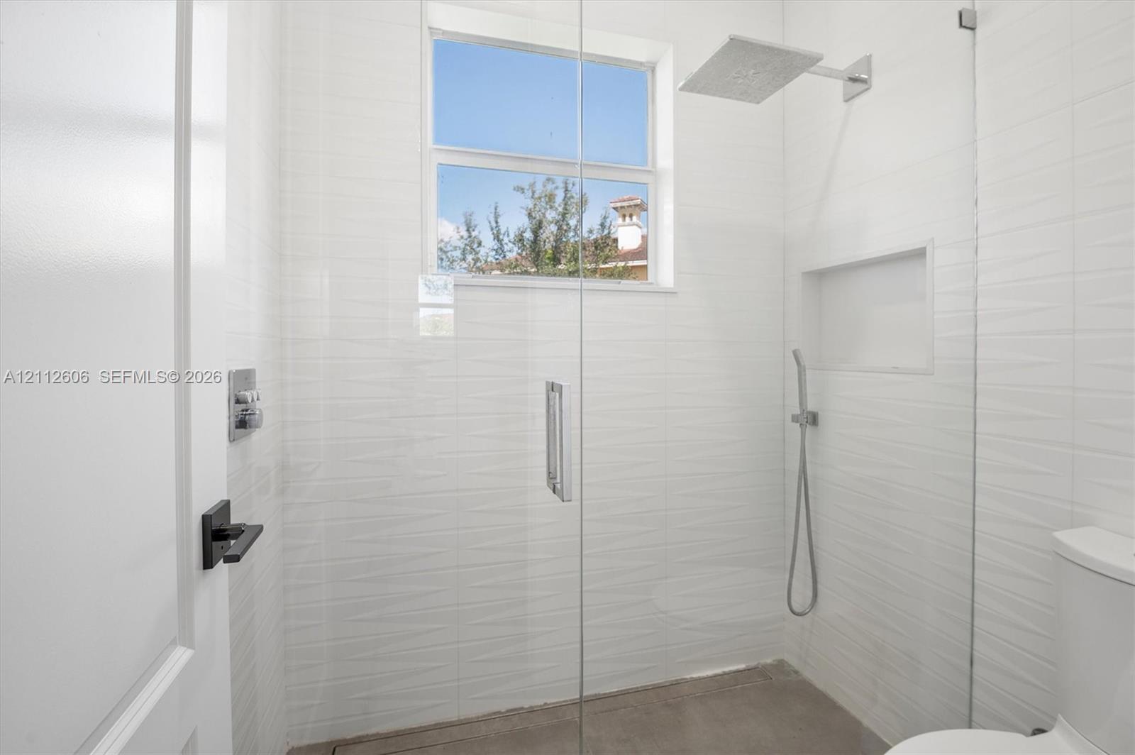 Walk-in shower featuring textured white wall tile, a frameless glass door, a rainfall showerhead, a hand-held sprayer, and an integrated wall niche