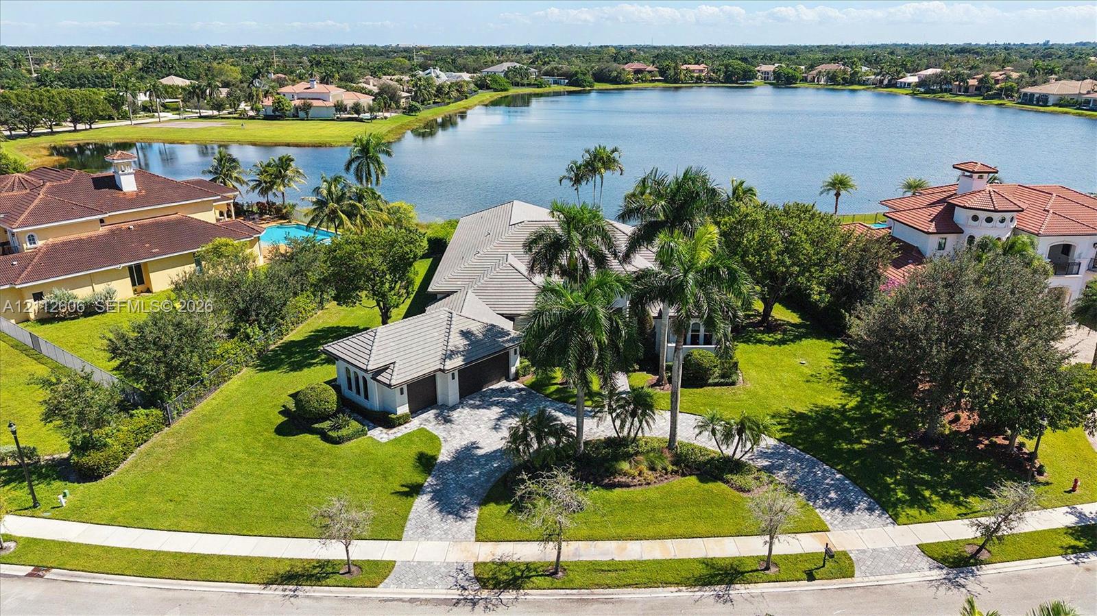 Aerial perspective featuring a multi-gabled residence with a concrete tile roof, a paved circular driveway, mature palm landscaping, and expansive water frontage