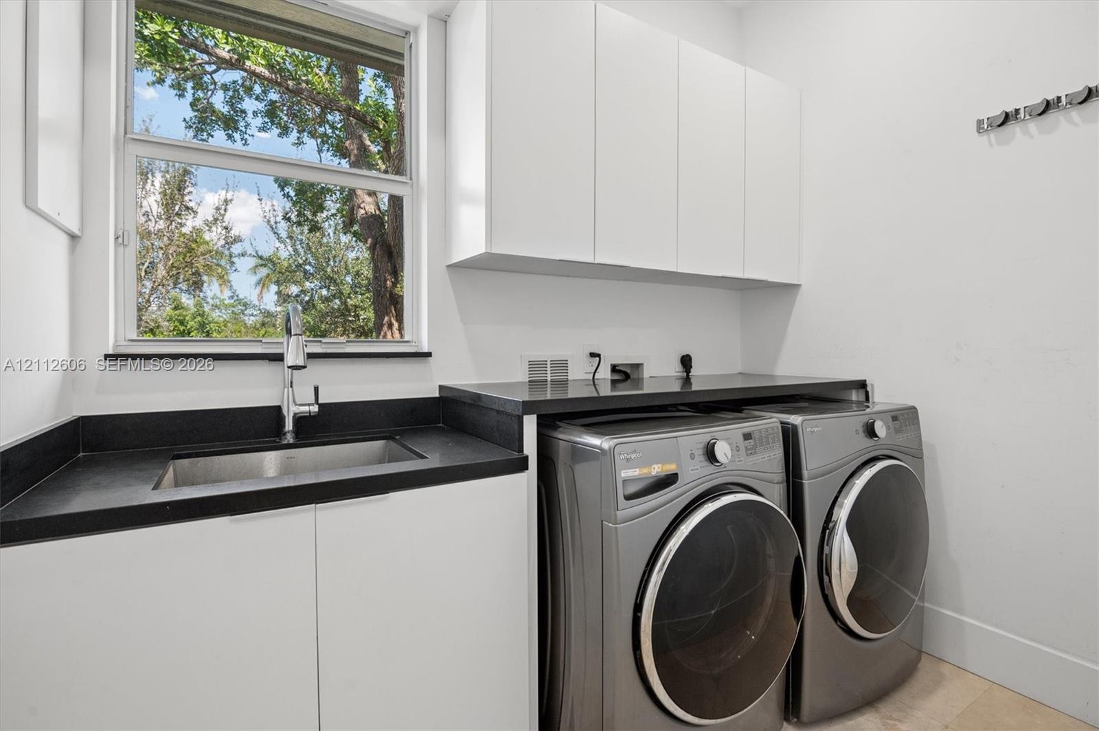 Laundry space featuring white cabinetry, dark solid-surface countertops, an undermount stainless steel sink with a chrome gooseneck faucet, and an exterior window