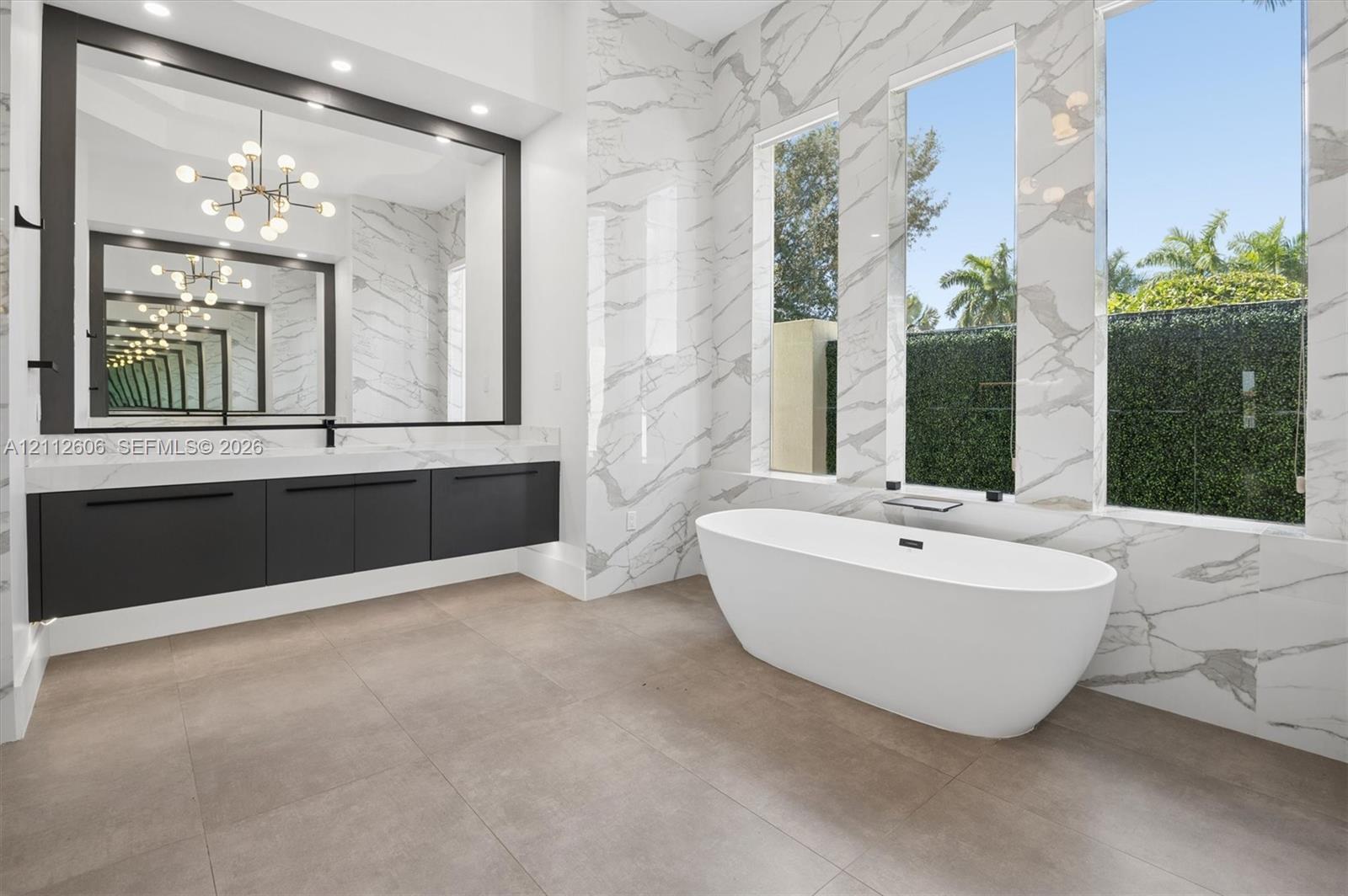 Bathroom featuring large-format tile flooring, a matte black floating vanity with a stone countertop, a freestanding soaking tub, floor-to-ceiling stone wall cladding, and oversized windows